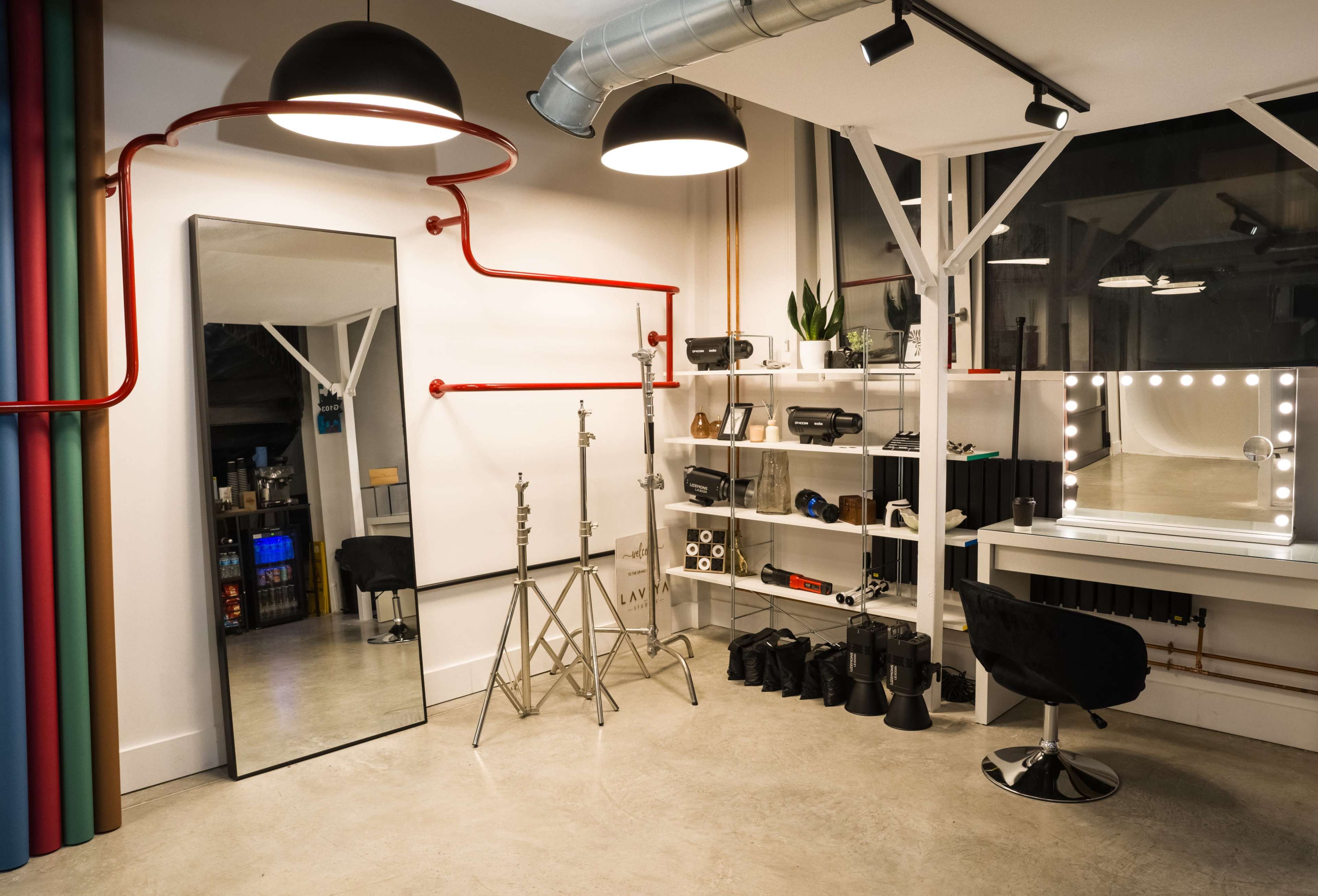 The image shows a modern studio space featuring a large mirror, lighting equipment on stands, and a makeup station with a lighted mirror, all set against a backdrop of colorful curtains and industrial piping.