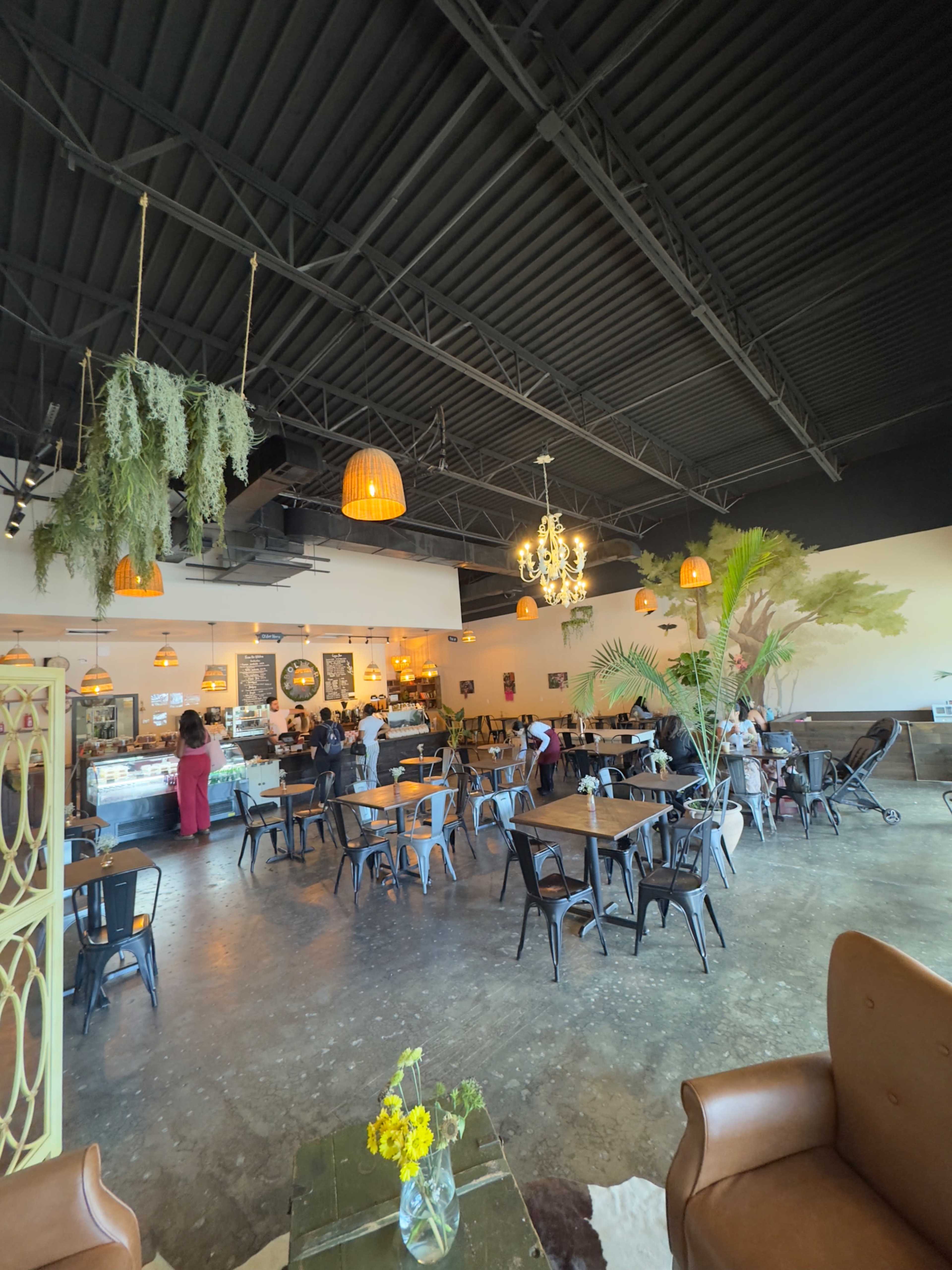 The image shows a spacious cafe interior featuring various tables and chairs, pendant lighting, and a small counter area with a display case.