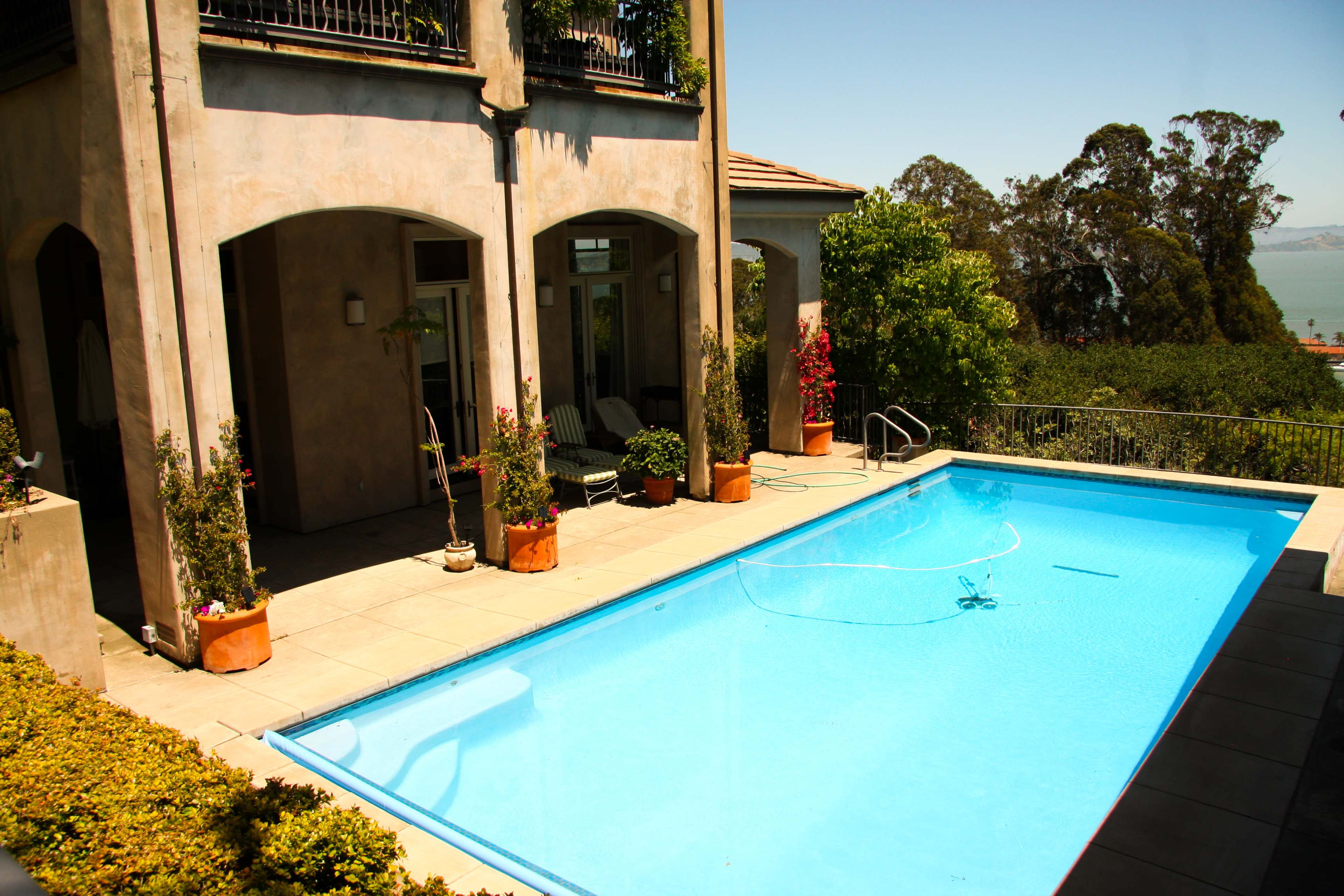 The image shows a residential backyard with a swimming pool, surrounded by stone tiles and landscaped greenery, and a two-story house in the background.