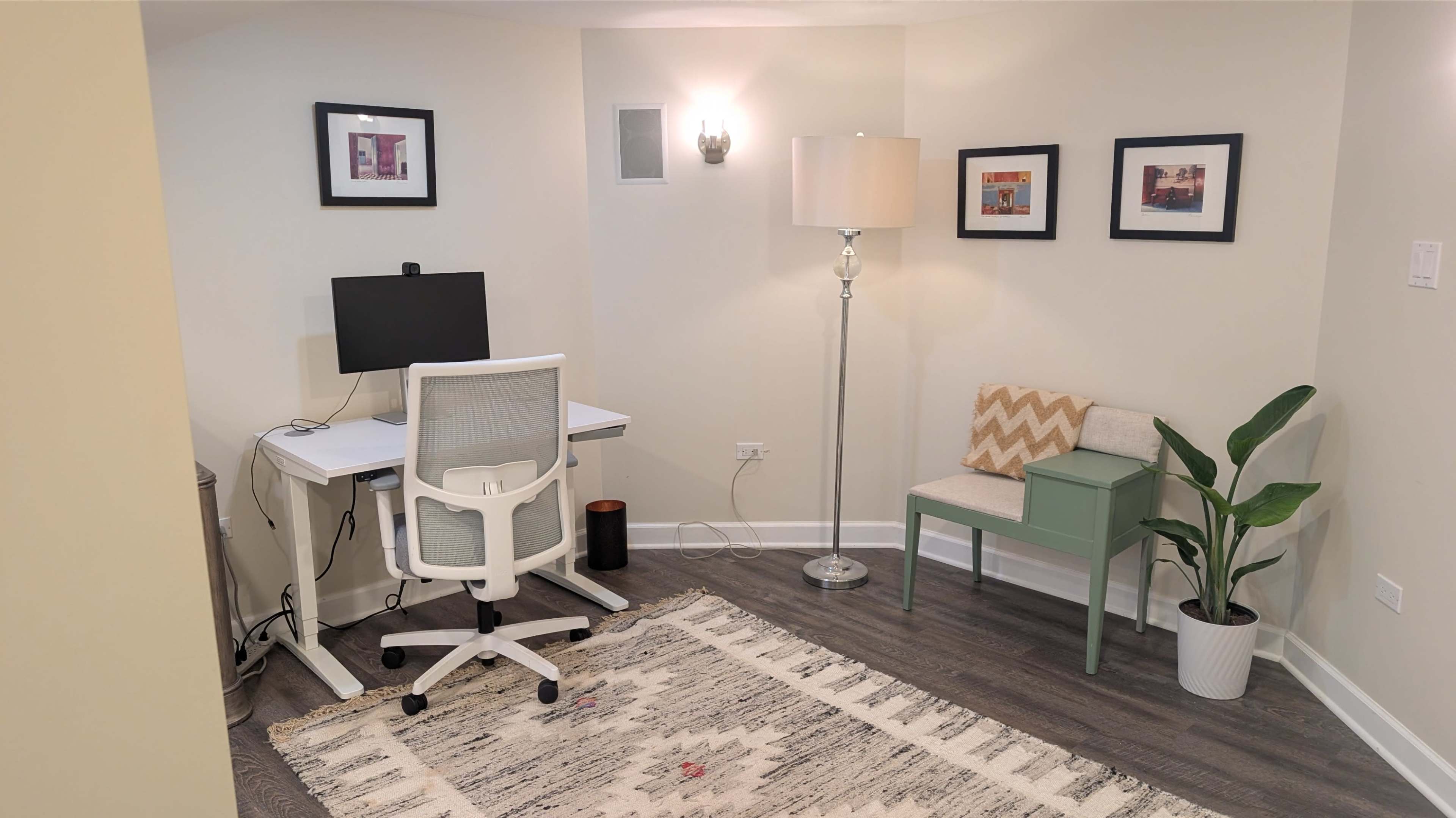 The image shows a small, well-lit home office space featuring a desk with a computer, a lamp, a chair, and a potted plant next to a decorative rug.