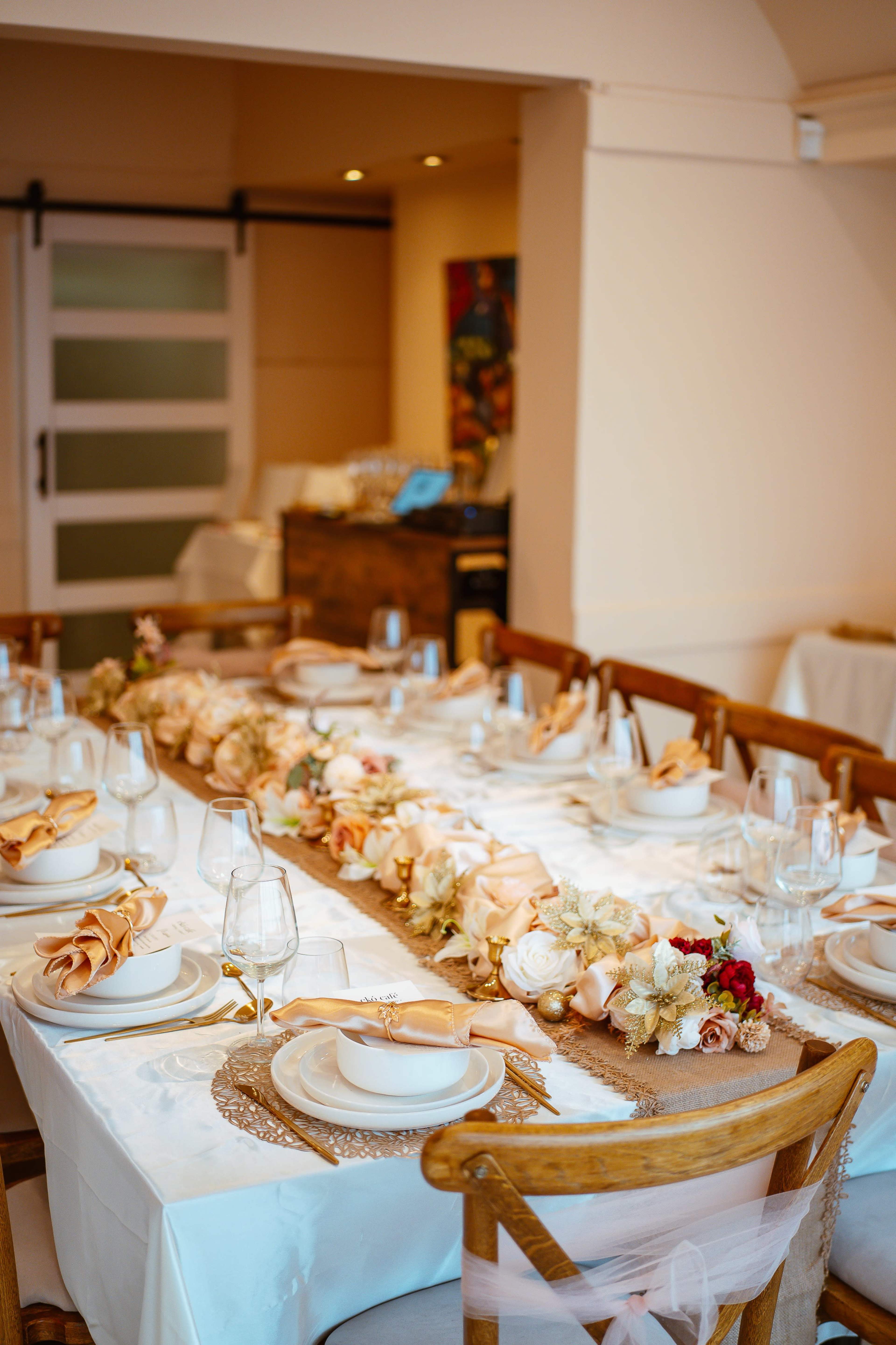 A dining table is elegantly set with white tablecloths, plates, glasses, and a floral centerpiece, arranged in a well-lit room.