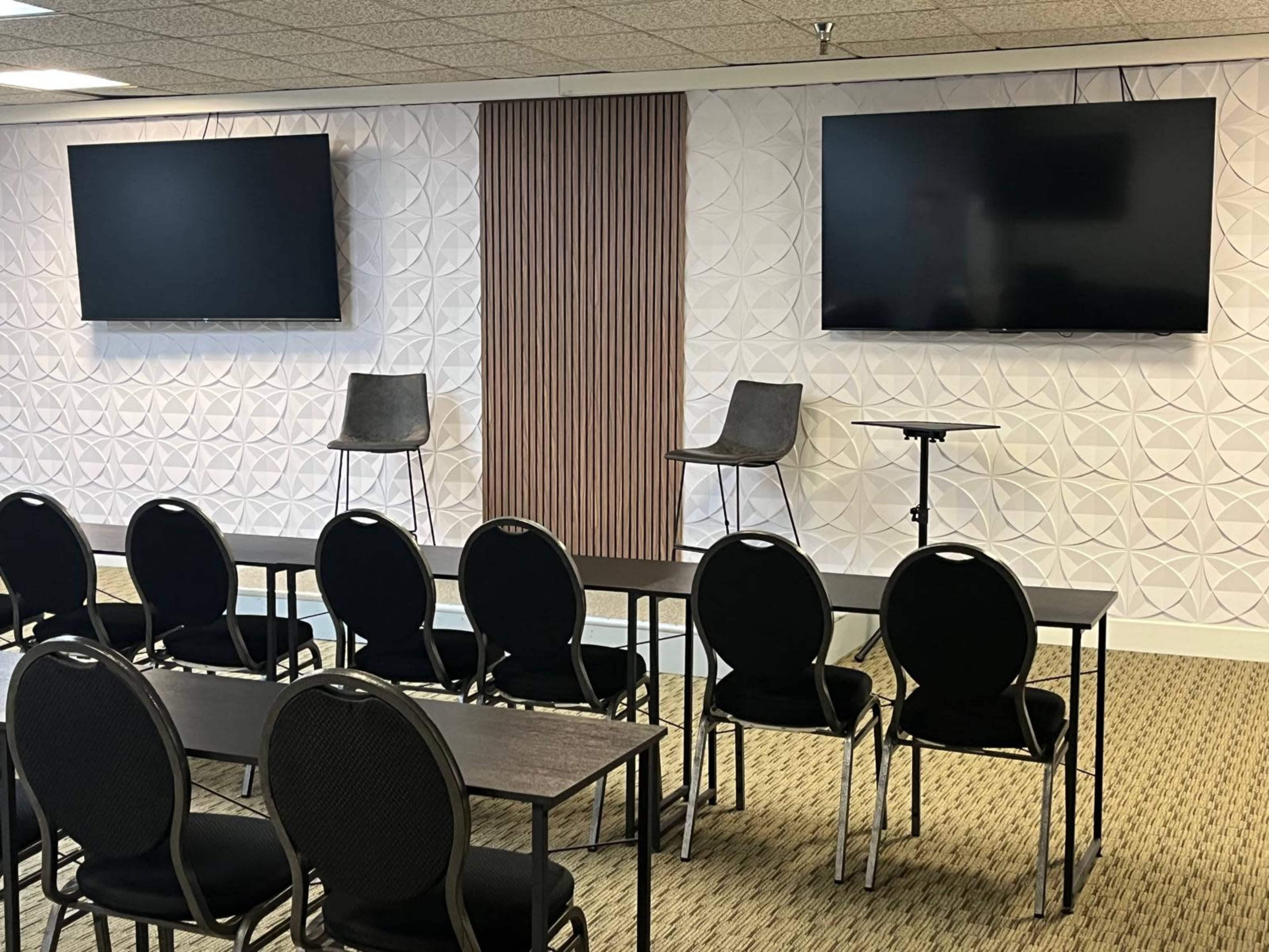 The image shows a conference room with two large screens mounted on a textured wall and several rows of black chairs facing a small table.