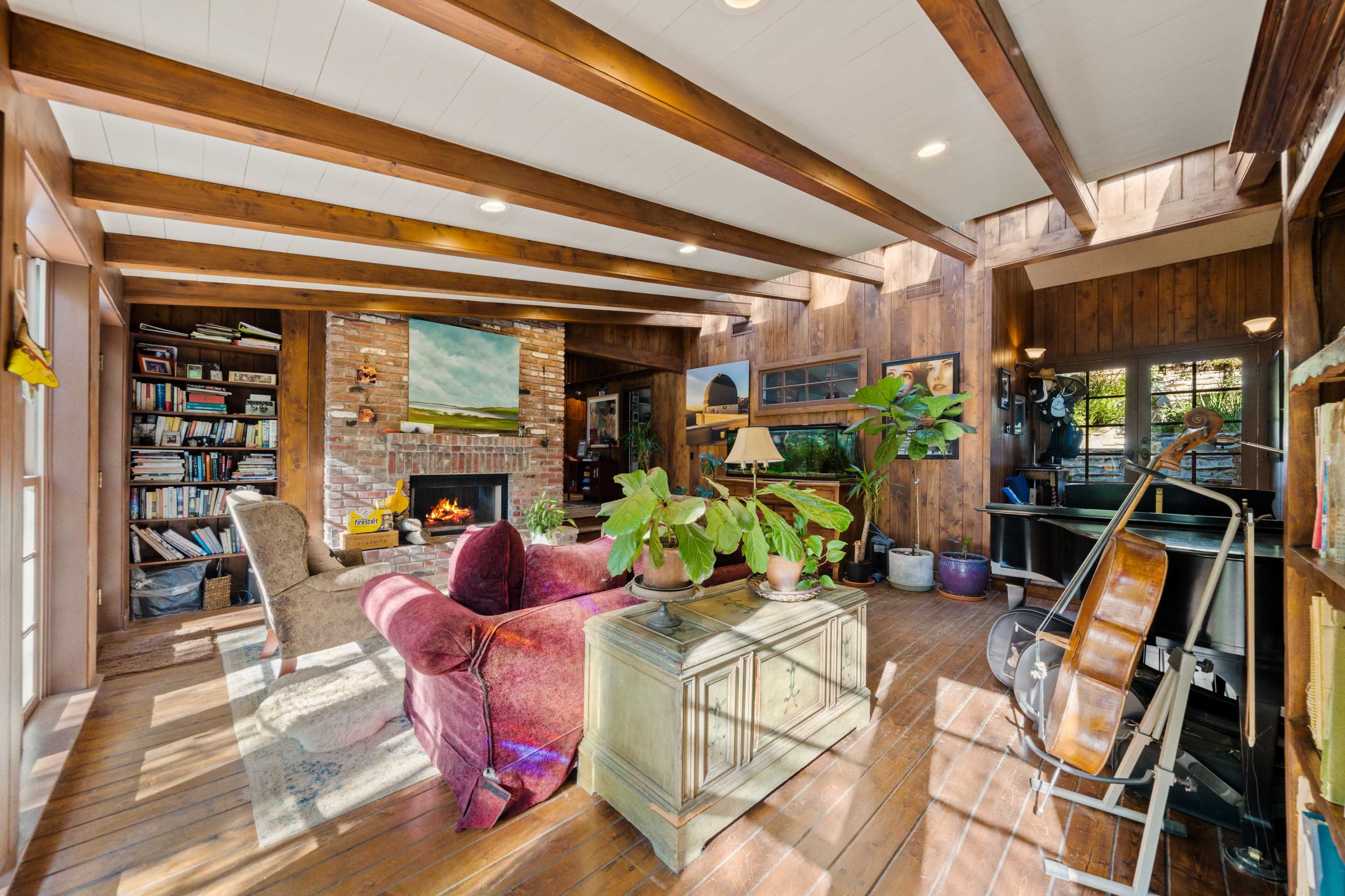 A cozy living room featuring a fireplace, wooden beams, bookshelves, potted plants, and a cello in the corner.