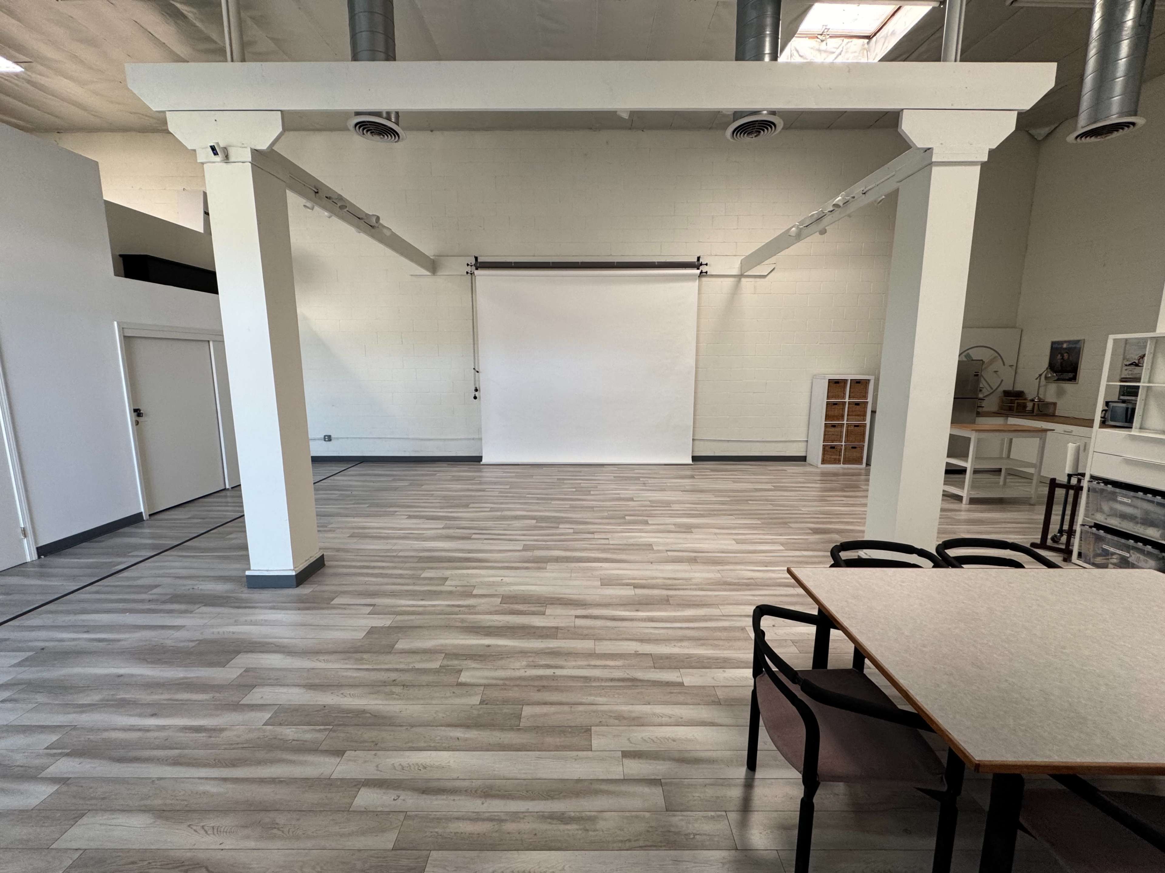 The image shows a spacious, empty studio with a white backdrop, wooden flooring, and a table with chairs in one corner.