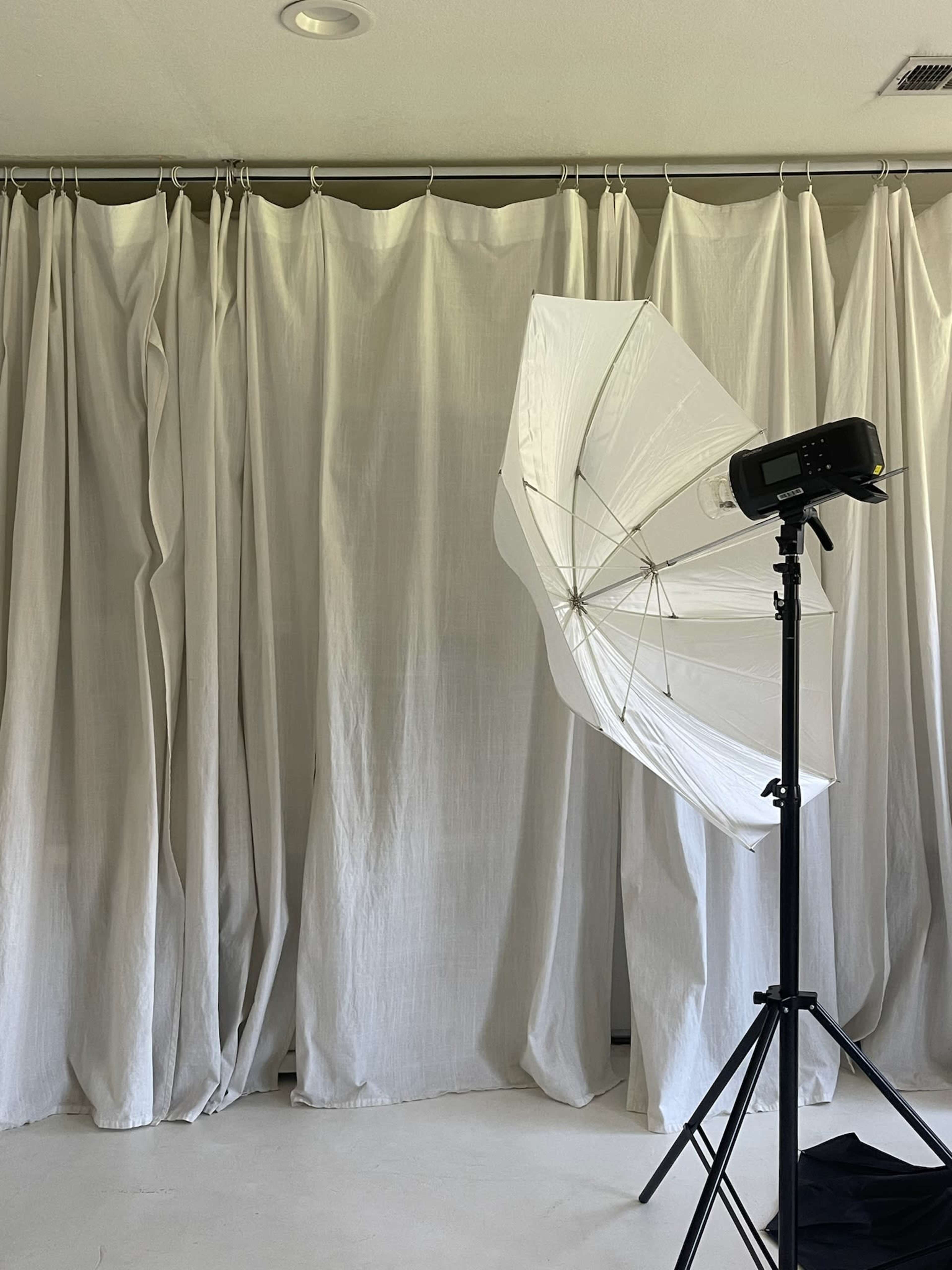 A photography setup features a large umbrella light positioned near a backdrop of draped white fabric curtains.