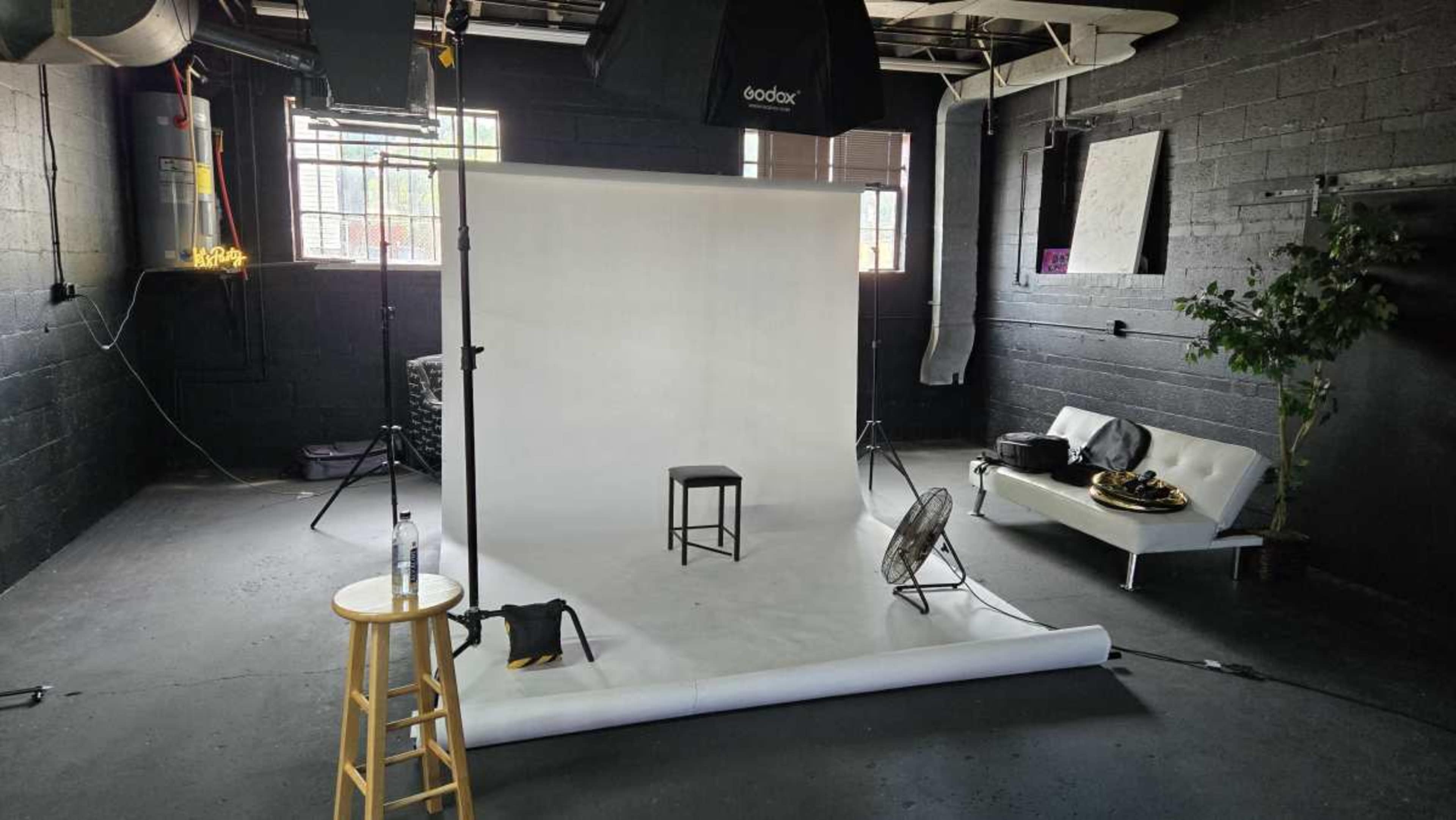 The image shows a photography studio with a white backdrop, a stool, a fan, and a couch against a black wall.