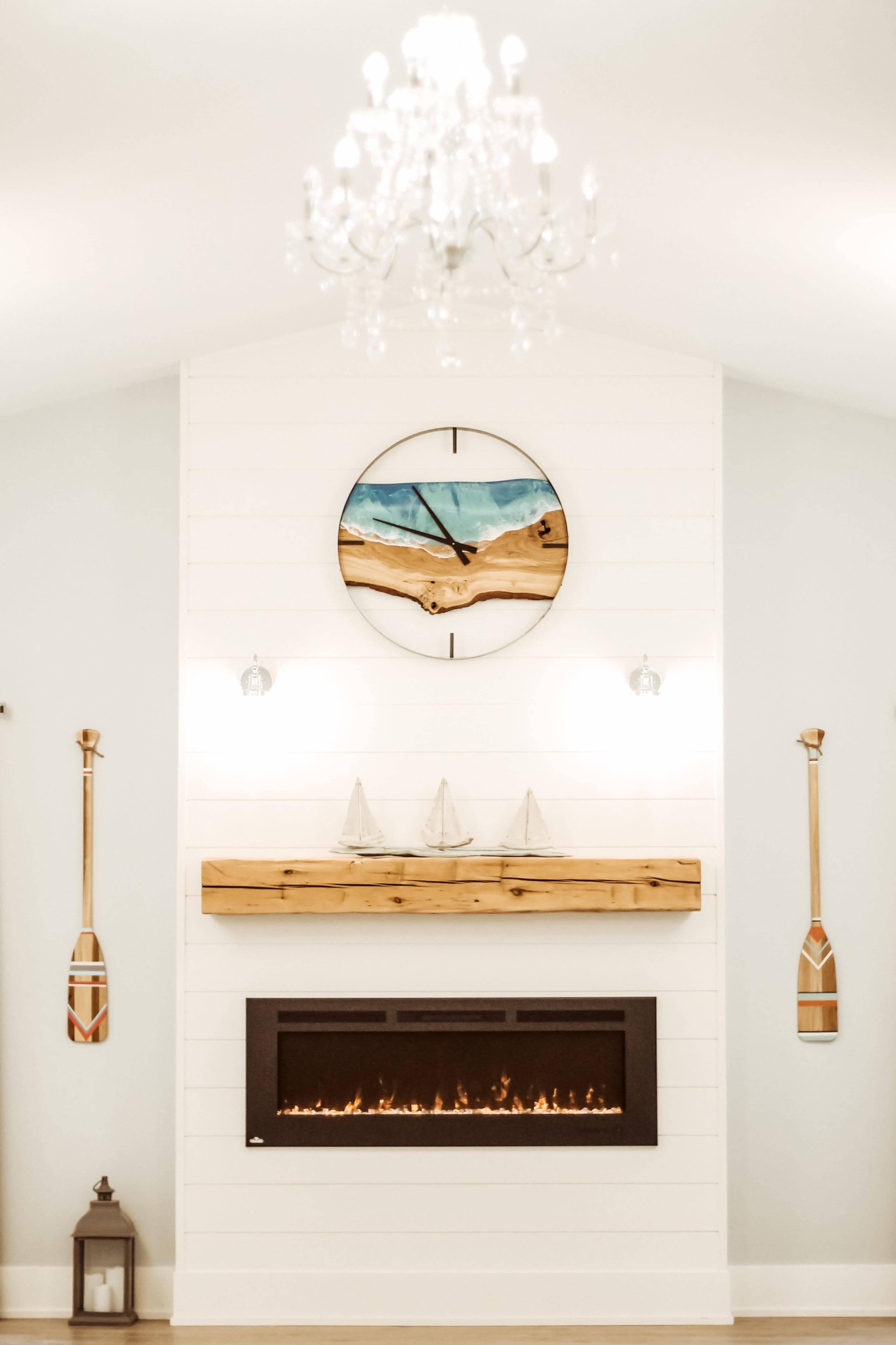 A modern living room with a white shiplap wall featuring a fireplace, a wooden mantel, decorative sailboat models, a round wall clock, and two paddle-shaped wall hangings, all topped by a chandelier.