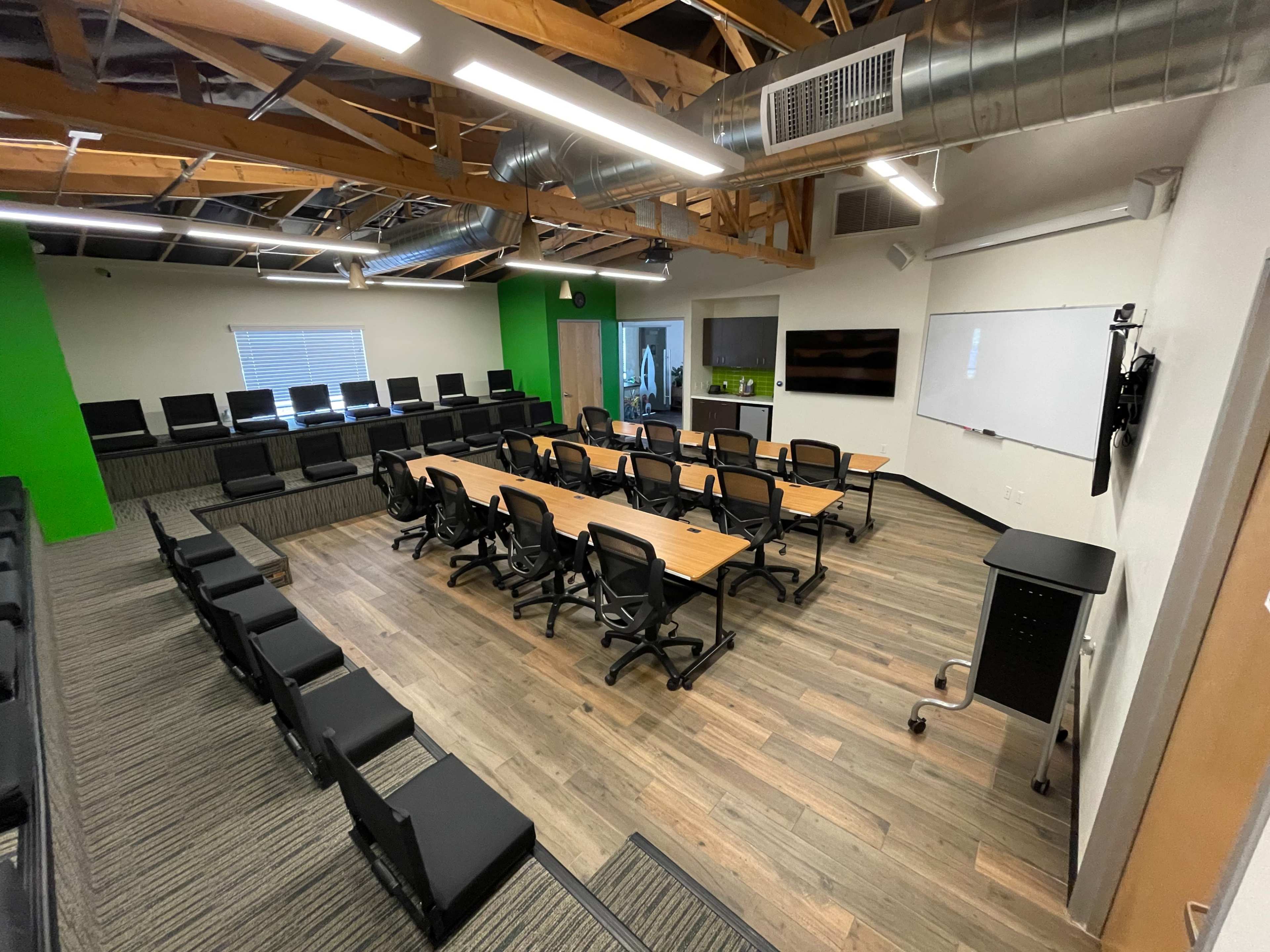 A modern conference room with a central table surrounded by black chairs, featuring a green accent wall and exposed wooden beams in the ceiling.