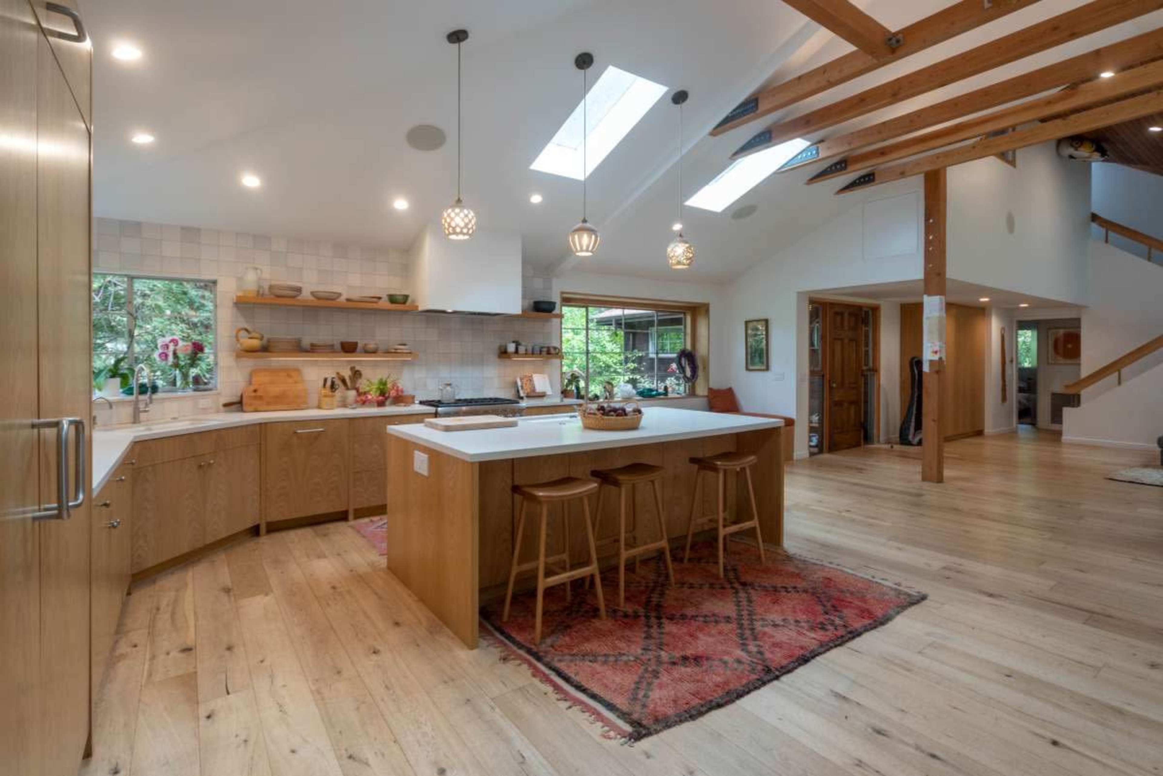 A modern kitchen features a large island with wooden cabinetry, pendant lights, and a view into a bright living space with wooden beams and a staircase.