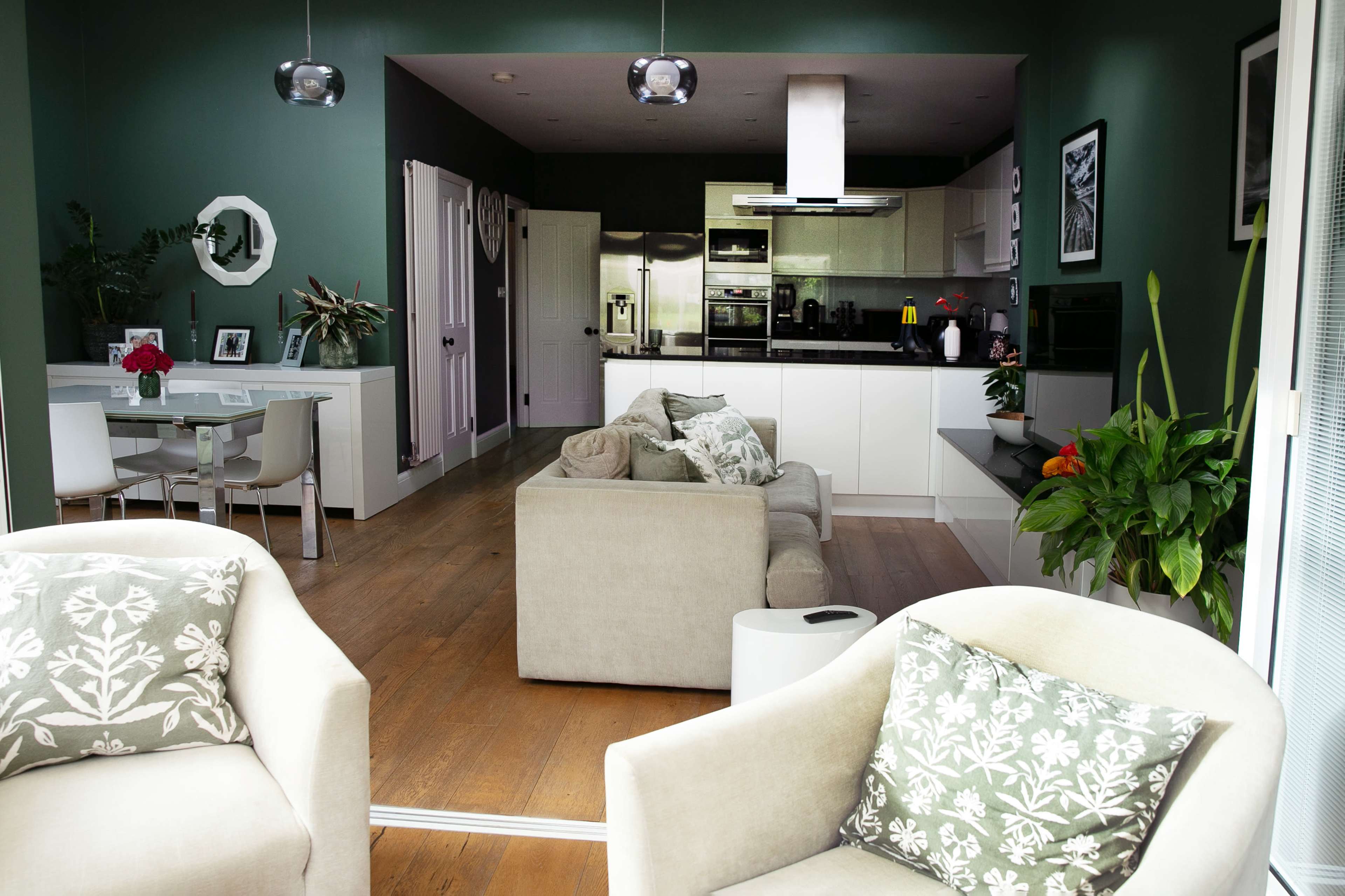 The image shows a modern open-plan living area with a kitchen, dining space, and seating area adorned with plants and decorative cushions.