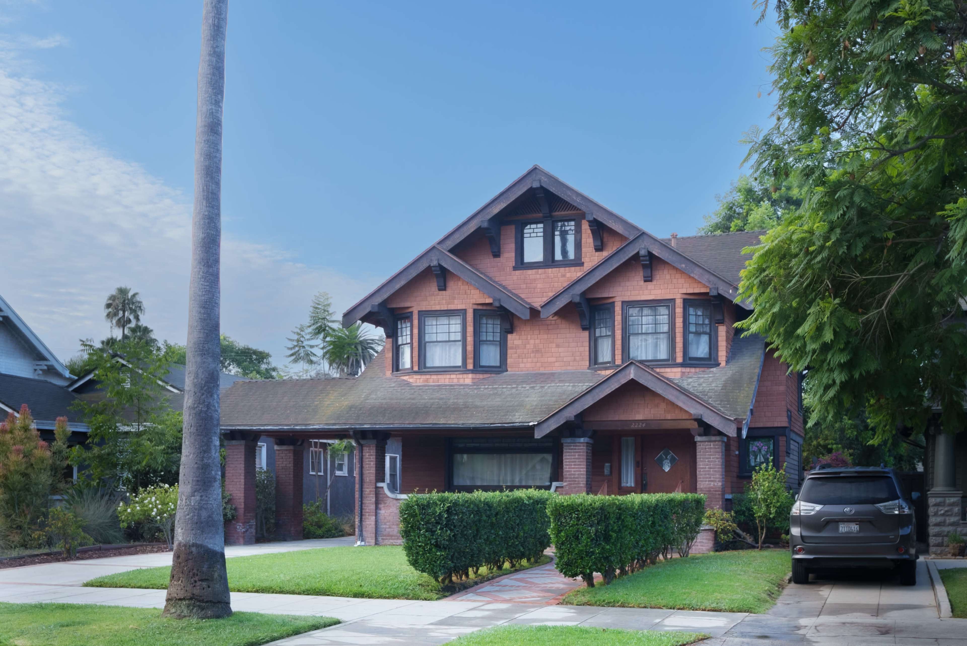 The image shows a two-story, craftsman-style house with a brick facade, surrounded by a manicured lawn and palm trees.