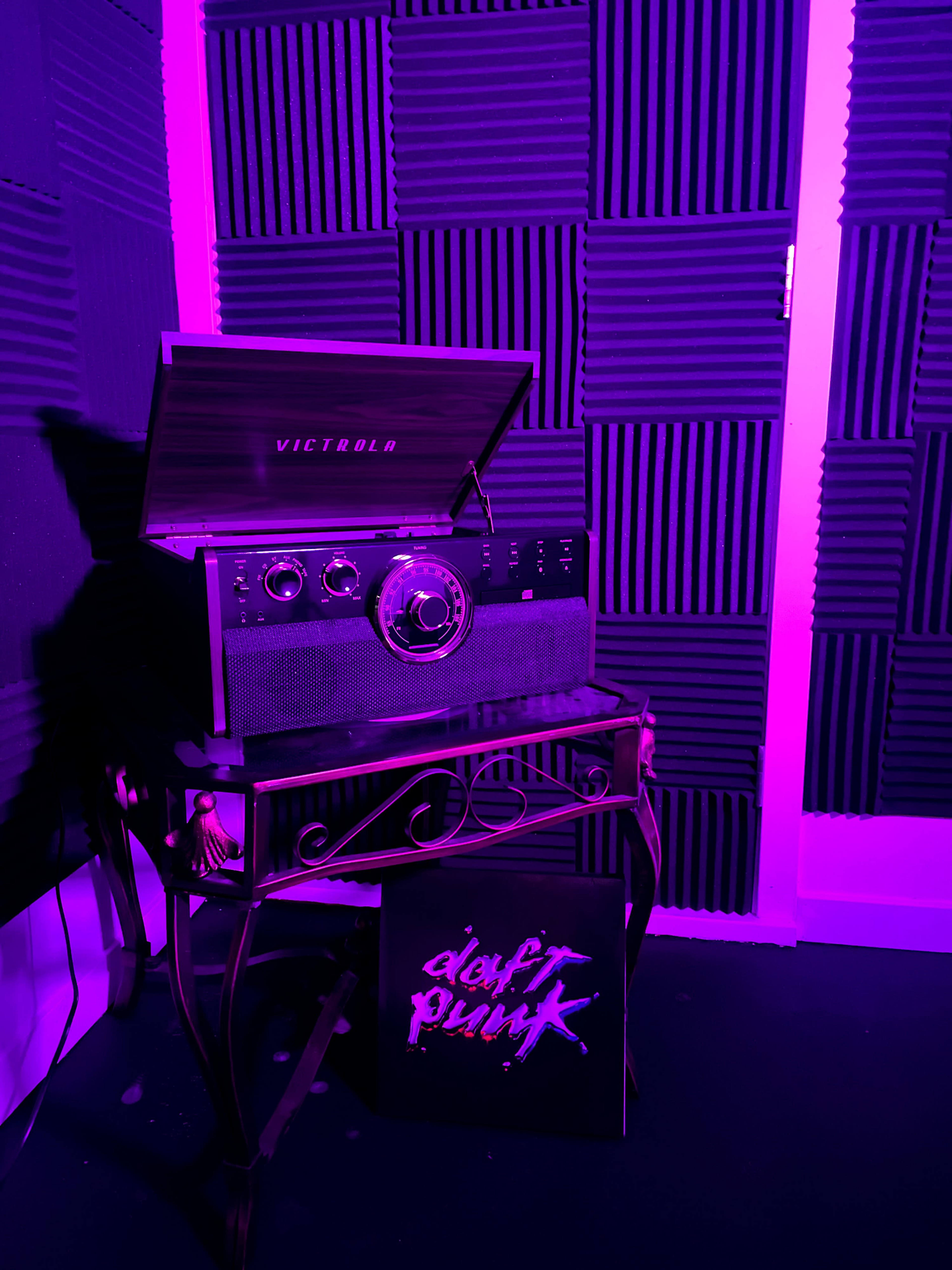 A vintage record player sits on a metal table beside a vinyl album cover featuring the text "Daft Punk," against a backdrop of acoustic foam panels.