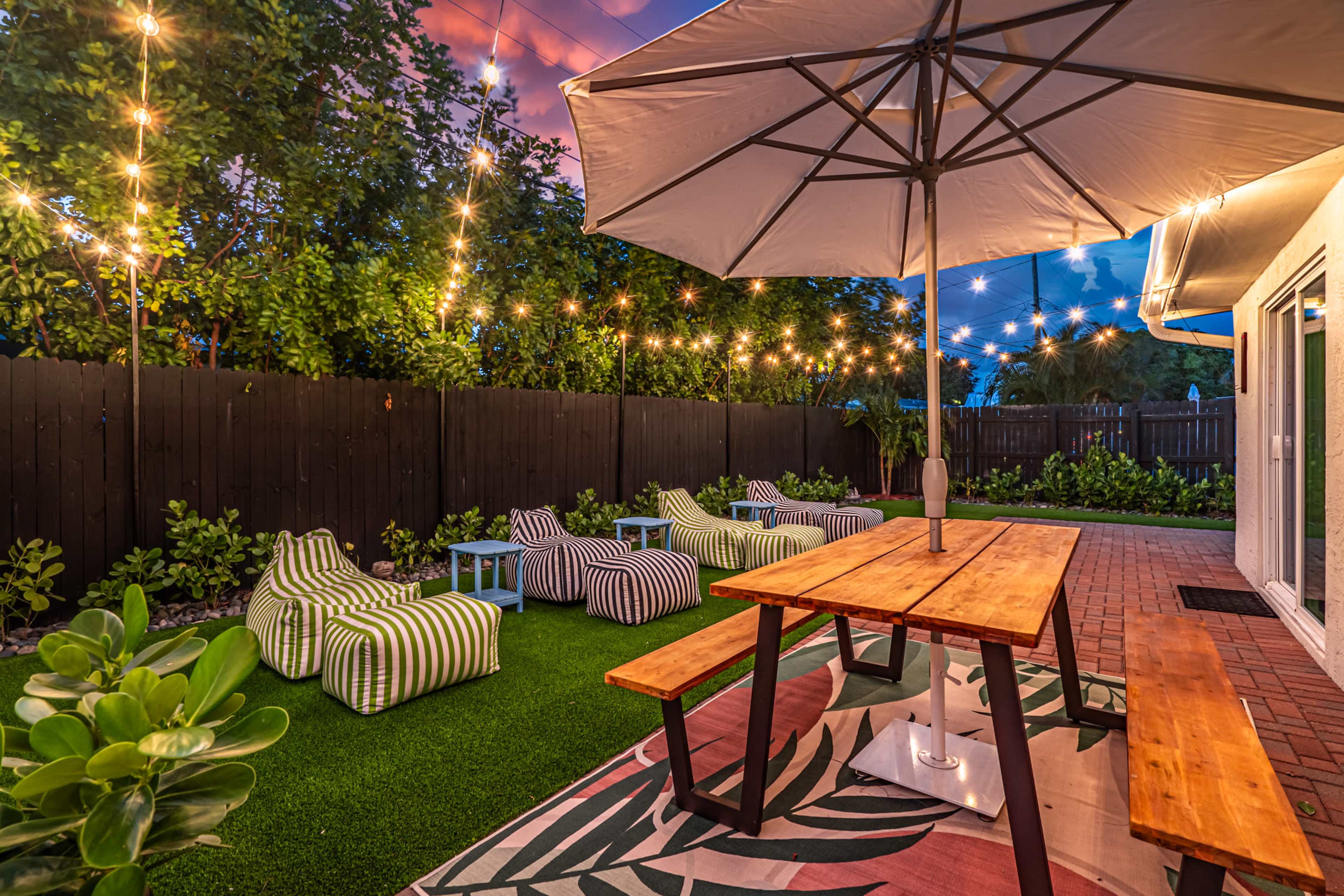 A cozy outdoor space features striped lounge chairs, a wooden dining table, and string lights hanging above, set against a backdrop of greenery and a colorful sunset sky.
