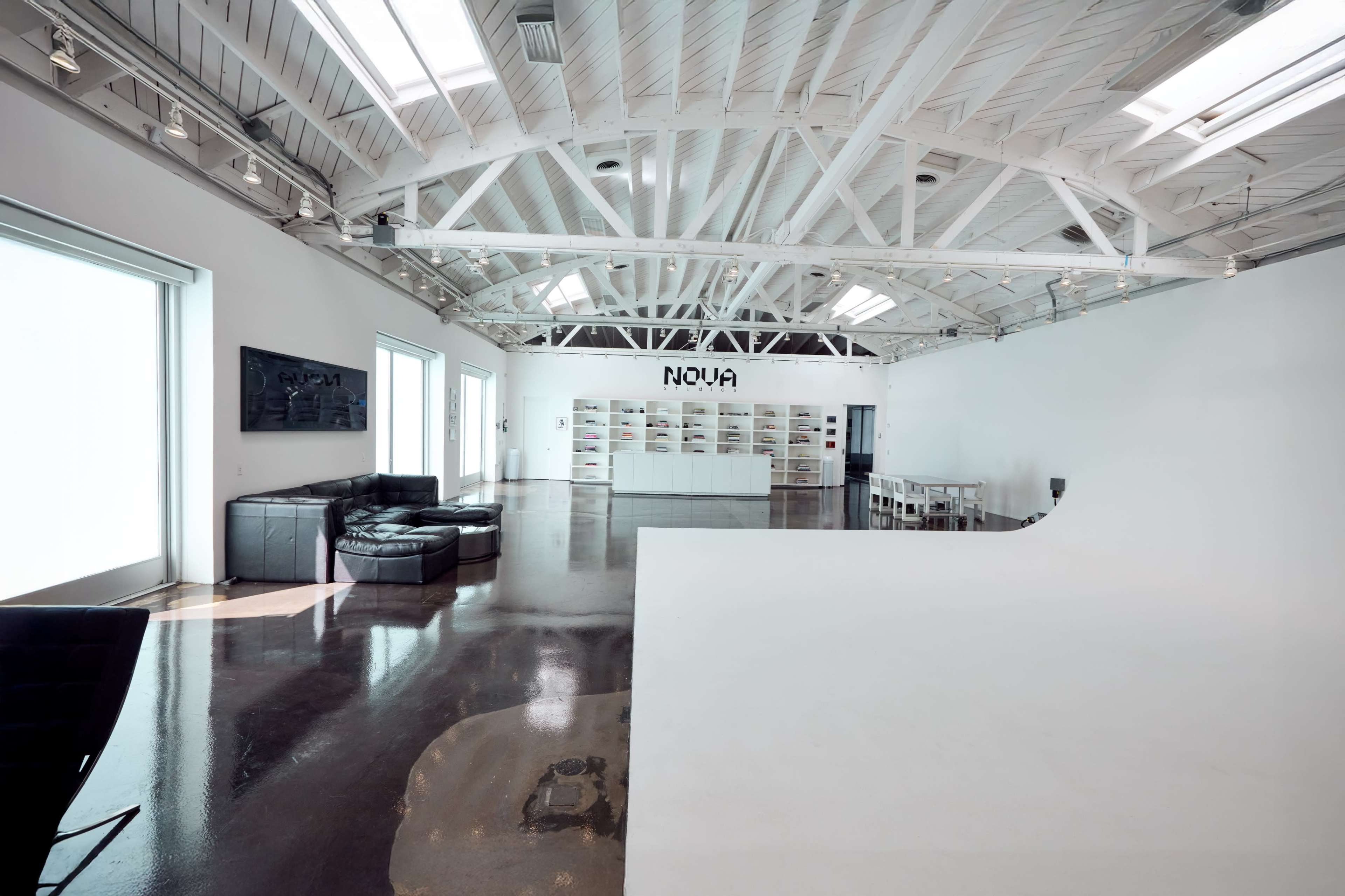 The image shows a spacious, well-lit interior with high ceilings, featuring a black sectional sofa, a white backdrop, and a shelf labeled "NOVA" against a stark white wall.