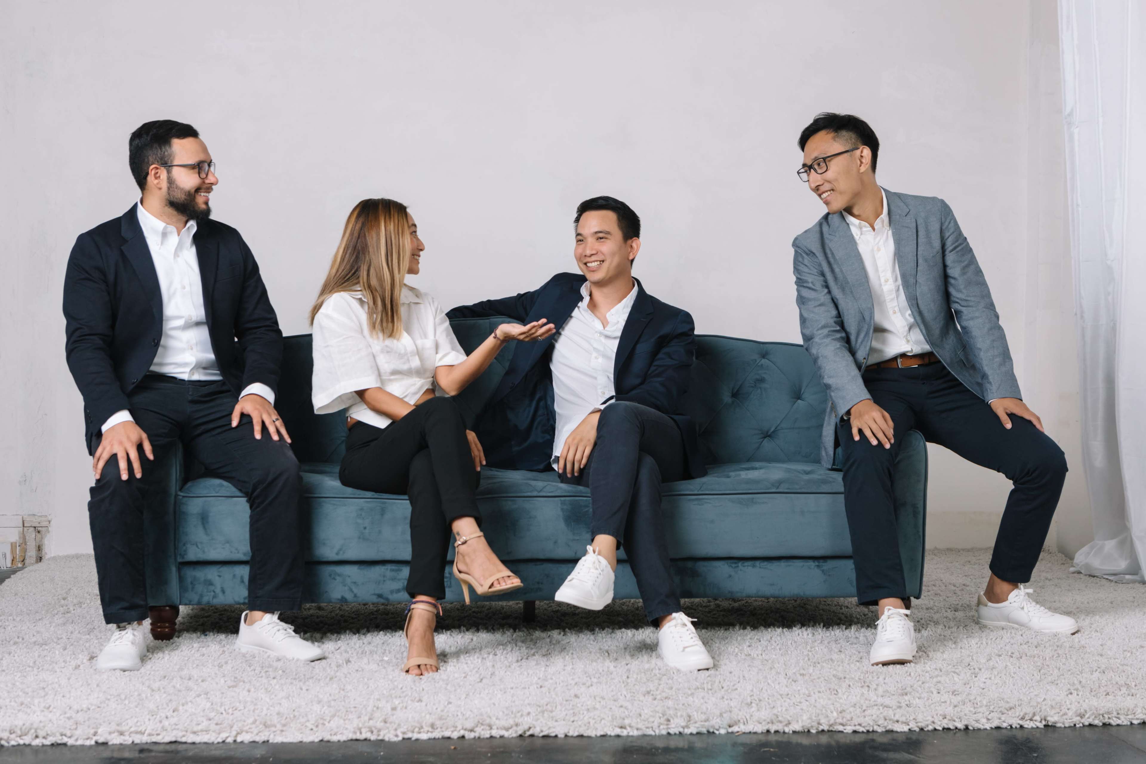 Four people are seated on a blue couch, engaged in conversation in a minimalist room with a white backdrop.