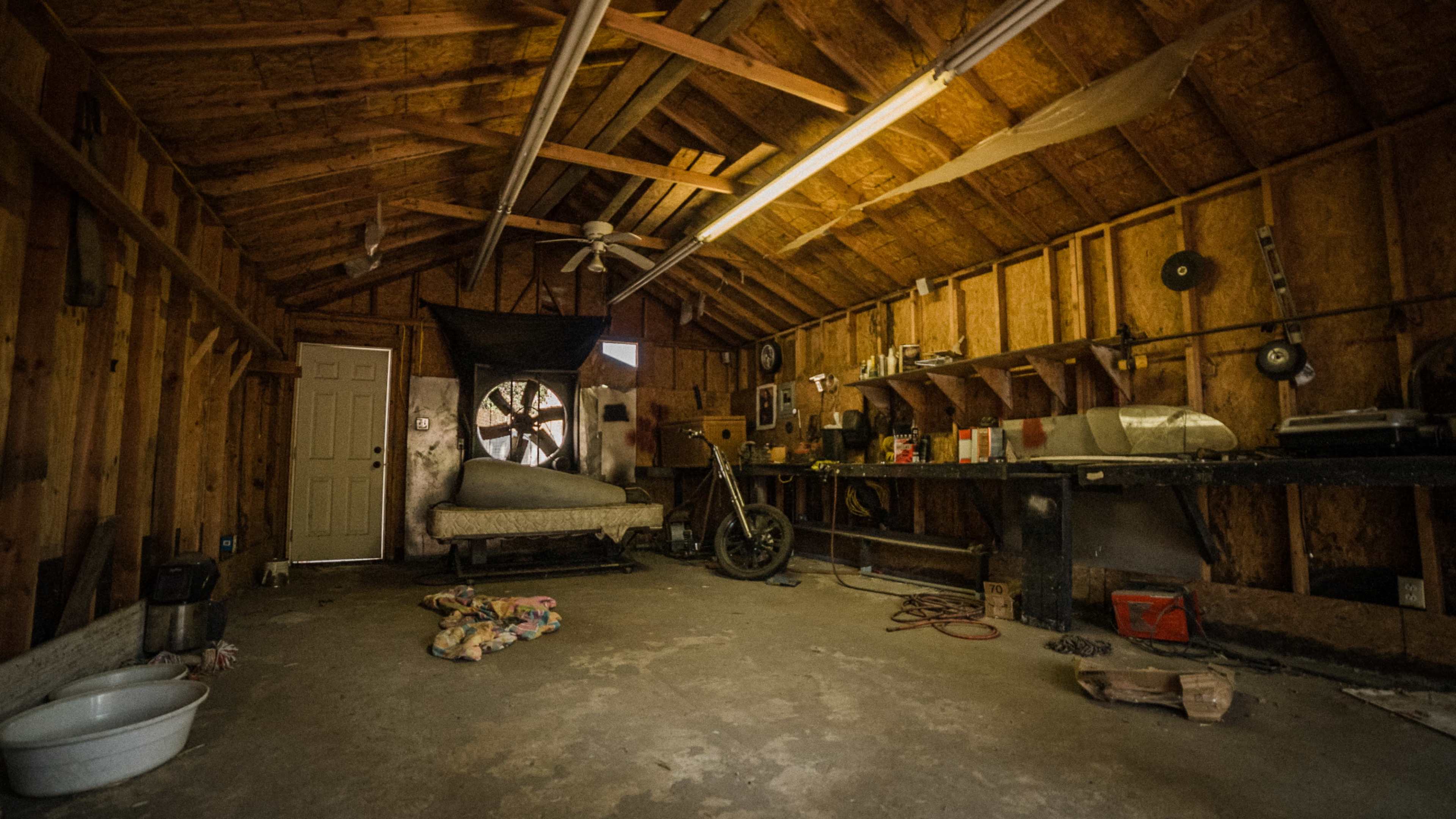The interior of a rustic garage features a bike, a workbench with tools, and a large fan mounted on the wall.