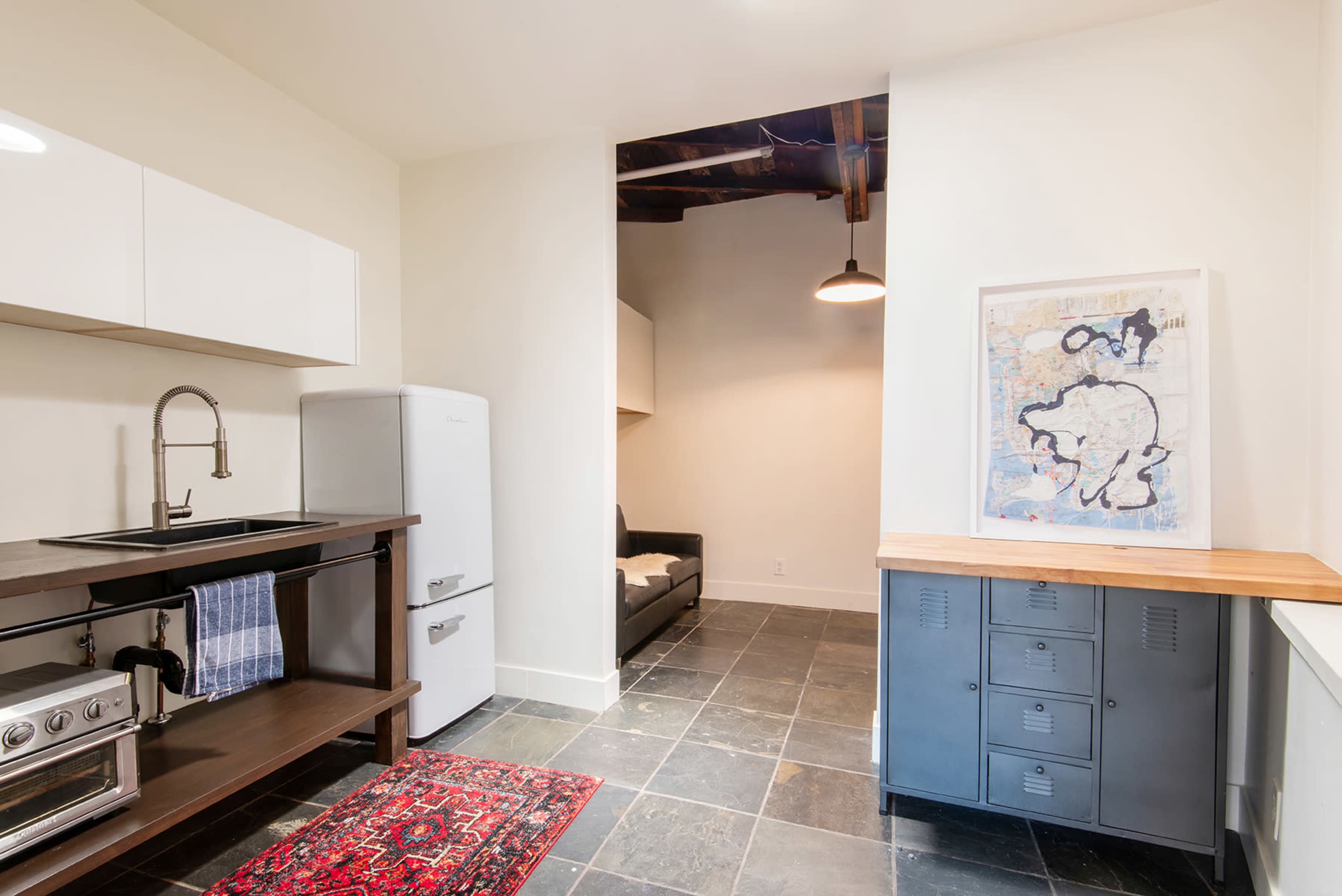 The image shows a kitchen with a sink and stove on one side, a white refrigerator, and a wooden table with metal lockers against a wall, illuminated by a pendant light.