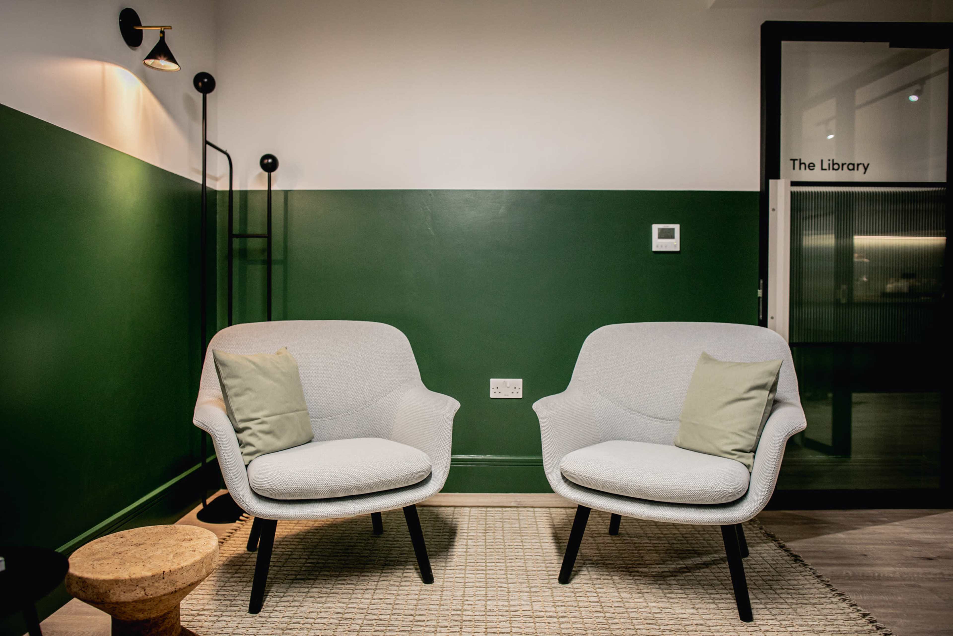 Two light gray chairs with green cushions are positioned opposite each other in a room with a dark green accent wall, beside a small wooden stool.