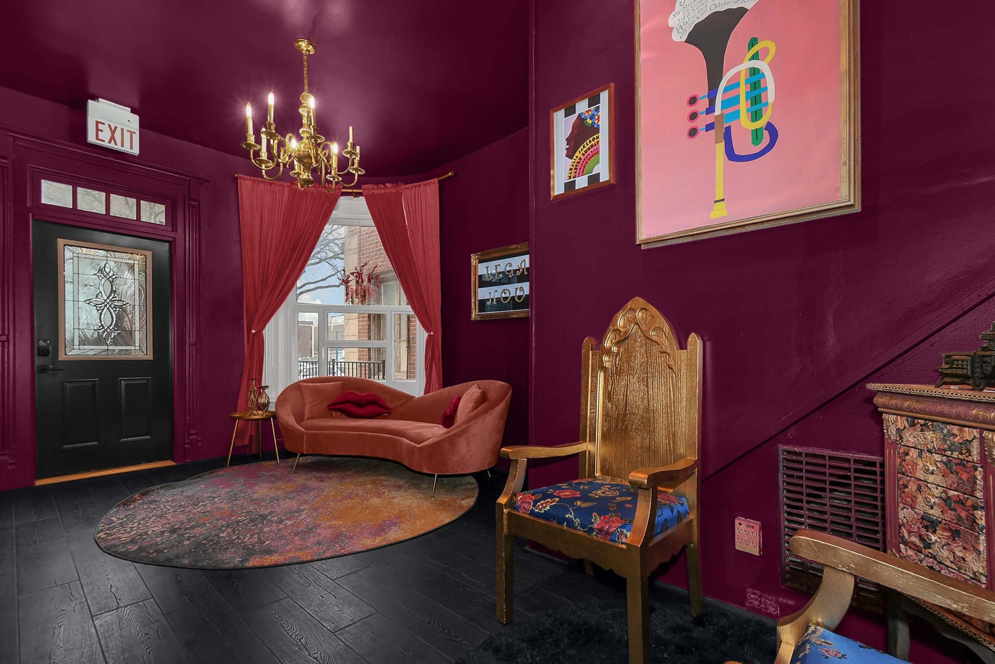 A small entryway featuring a velvet couch, a decorative gold chandelier, and a vintage armchair, all set against dark maroon walls.