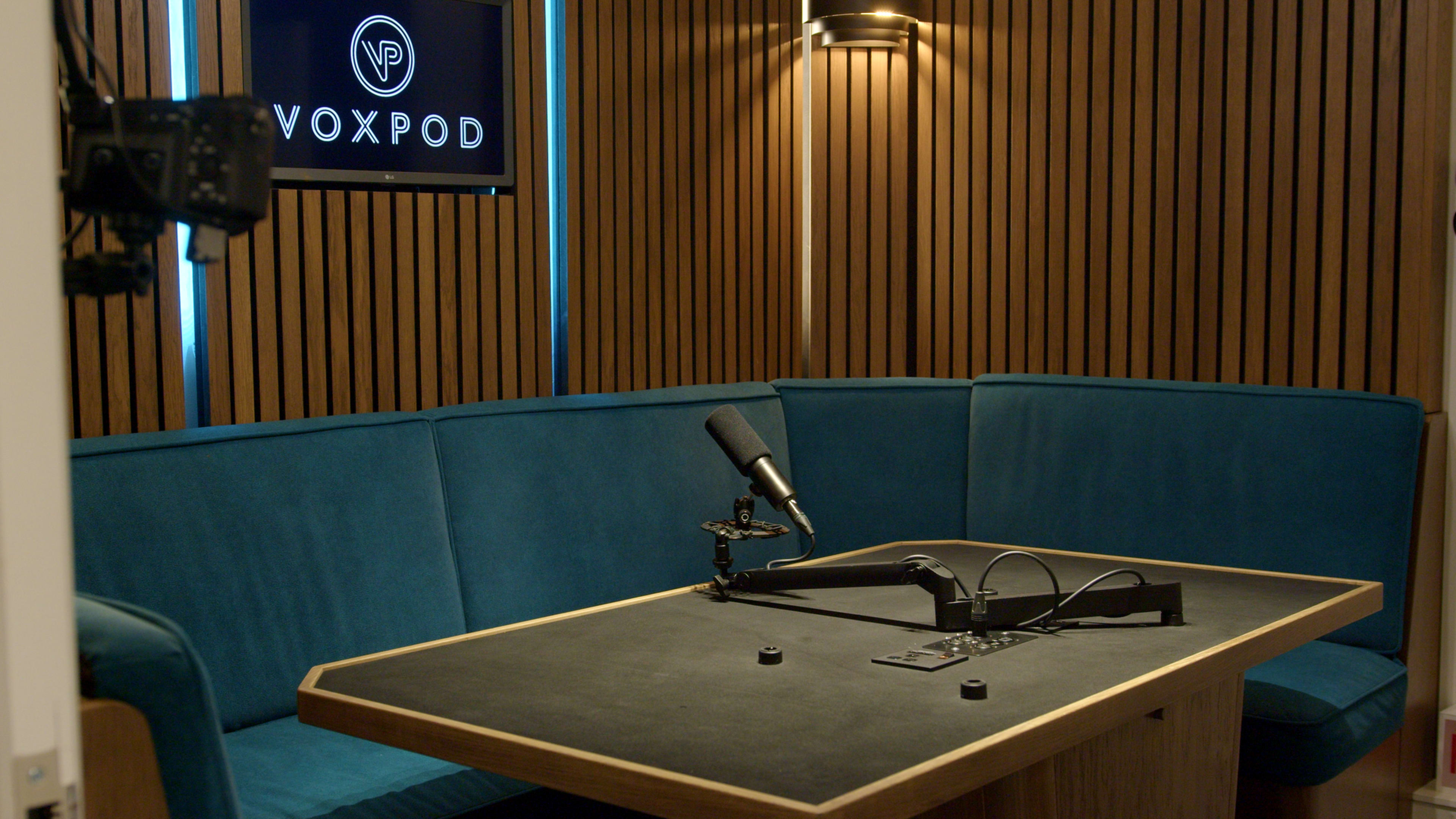 The image shows a small podcast recording studio featuring a large table with a microphone and a blue upholstered seating area against wooden paneling.