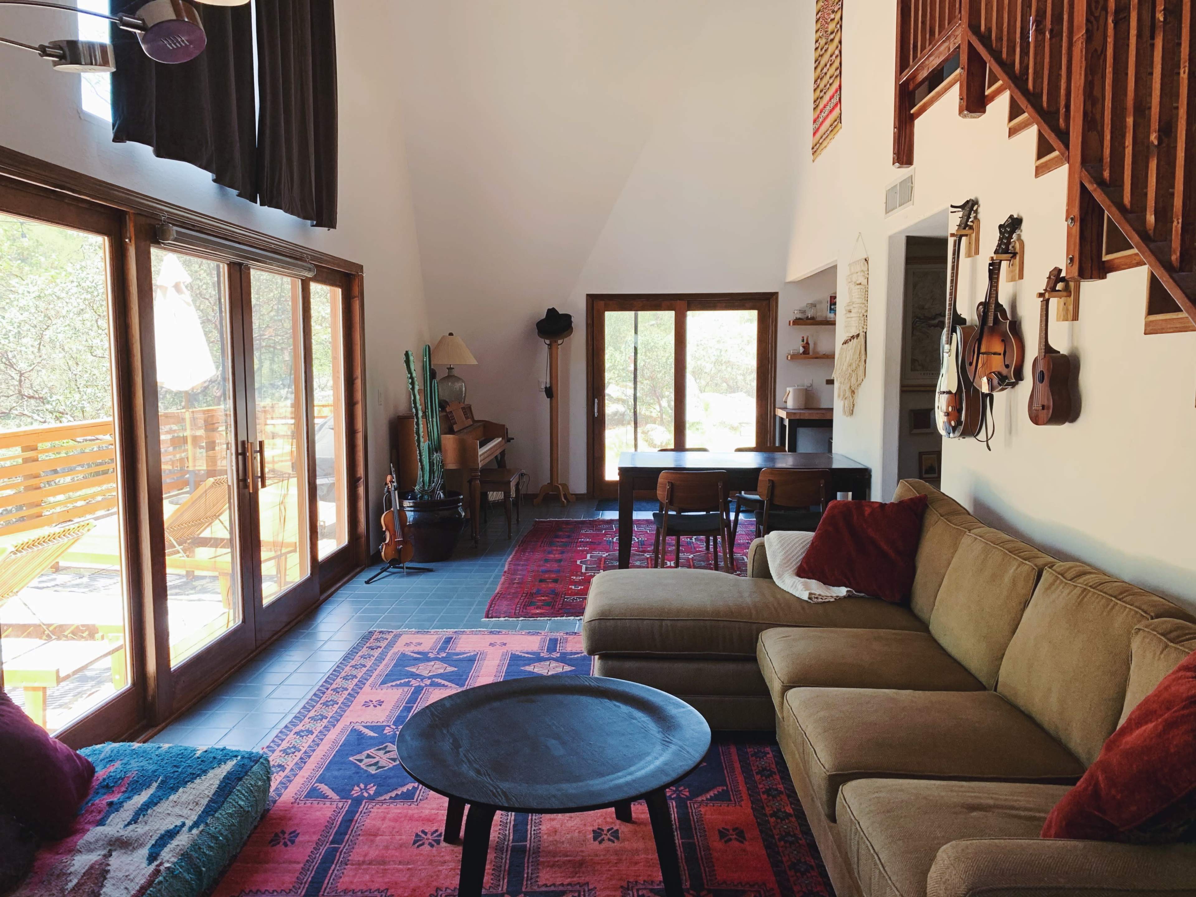 The interior of a modern living room features a large, beige sectional sofa, a round black coffee table, and large windows overlooking a patio, with wooden accents and various musical instruments displayed on the walls.