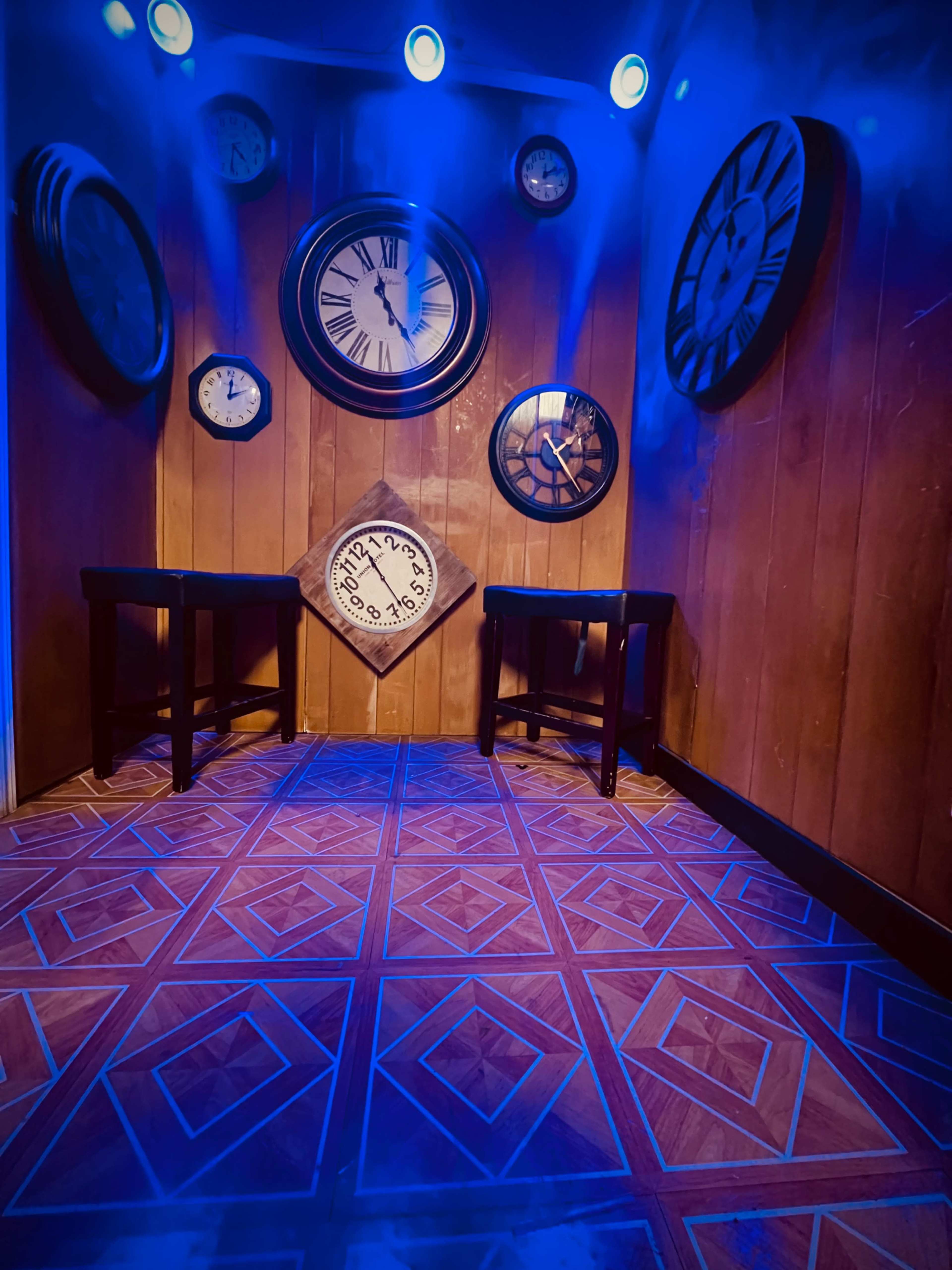 The image shows a dimly lit room with various wall clocks mounted on wooden walls and two small tables on the floor.