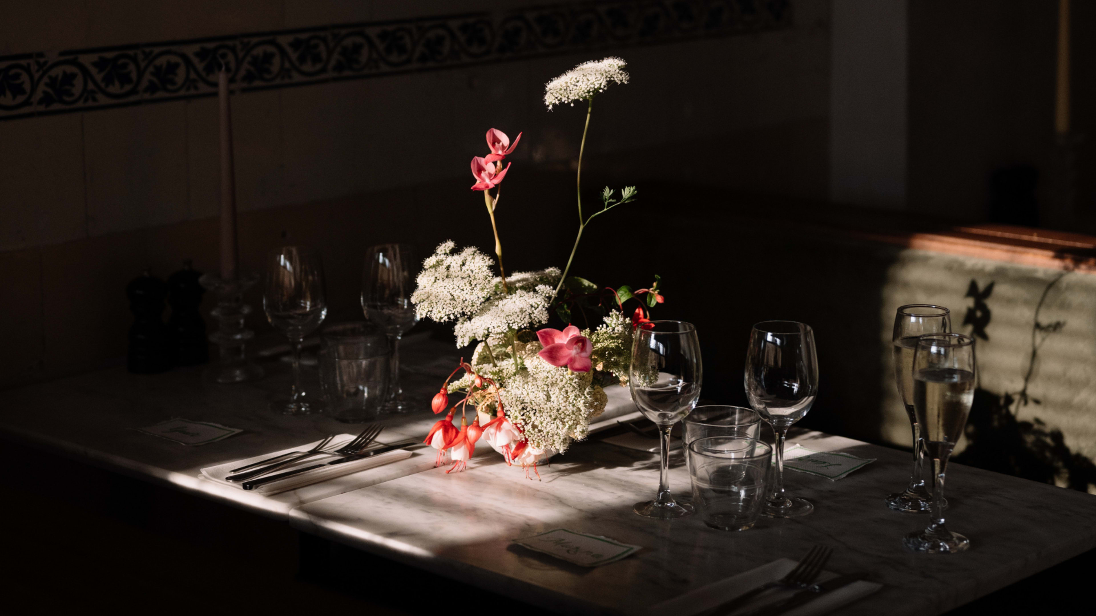 A dining table is set with glassware and silverware, featuring a floral arrangement at the center illuminated by sunlight.