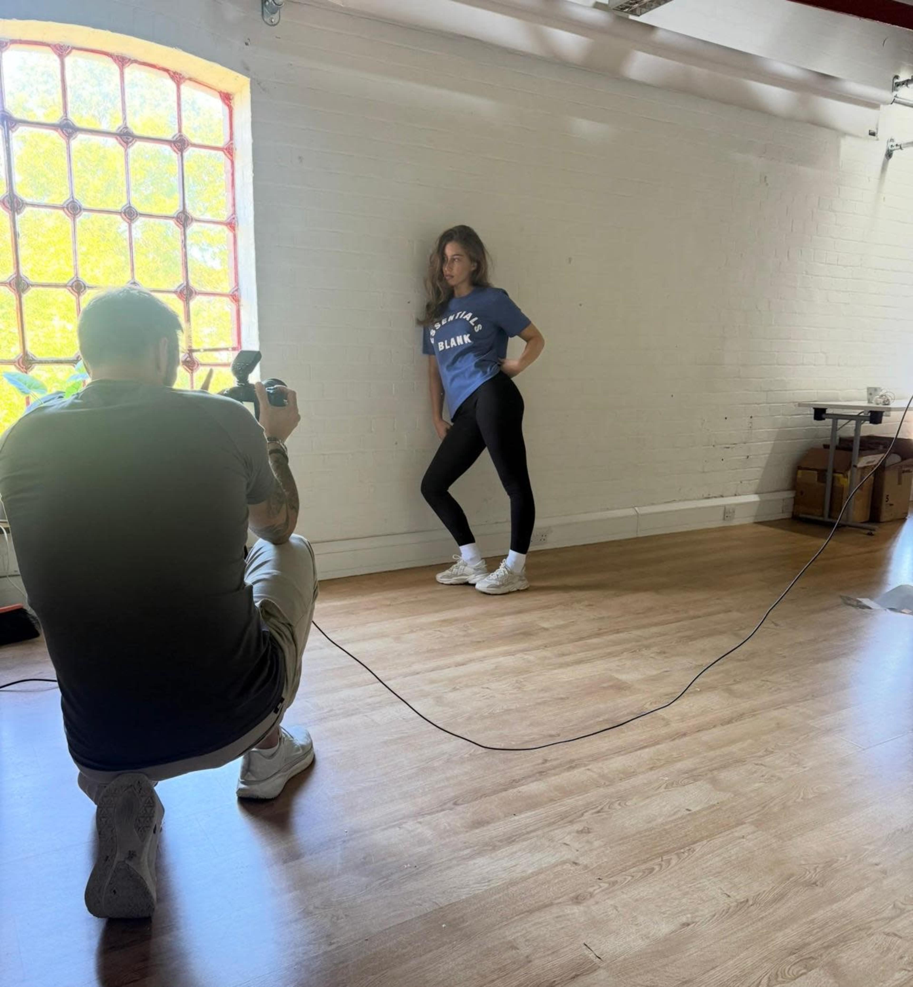 A photographer captures a model posing against a wall in a well-lit studio.