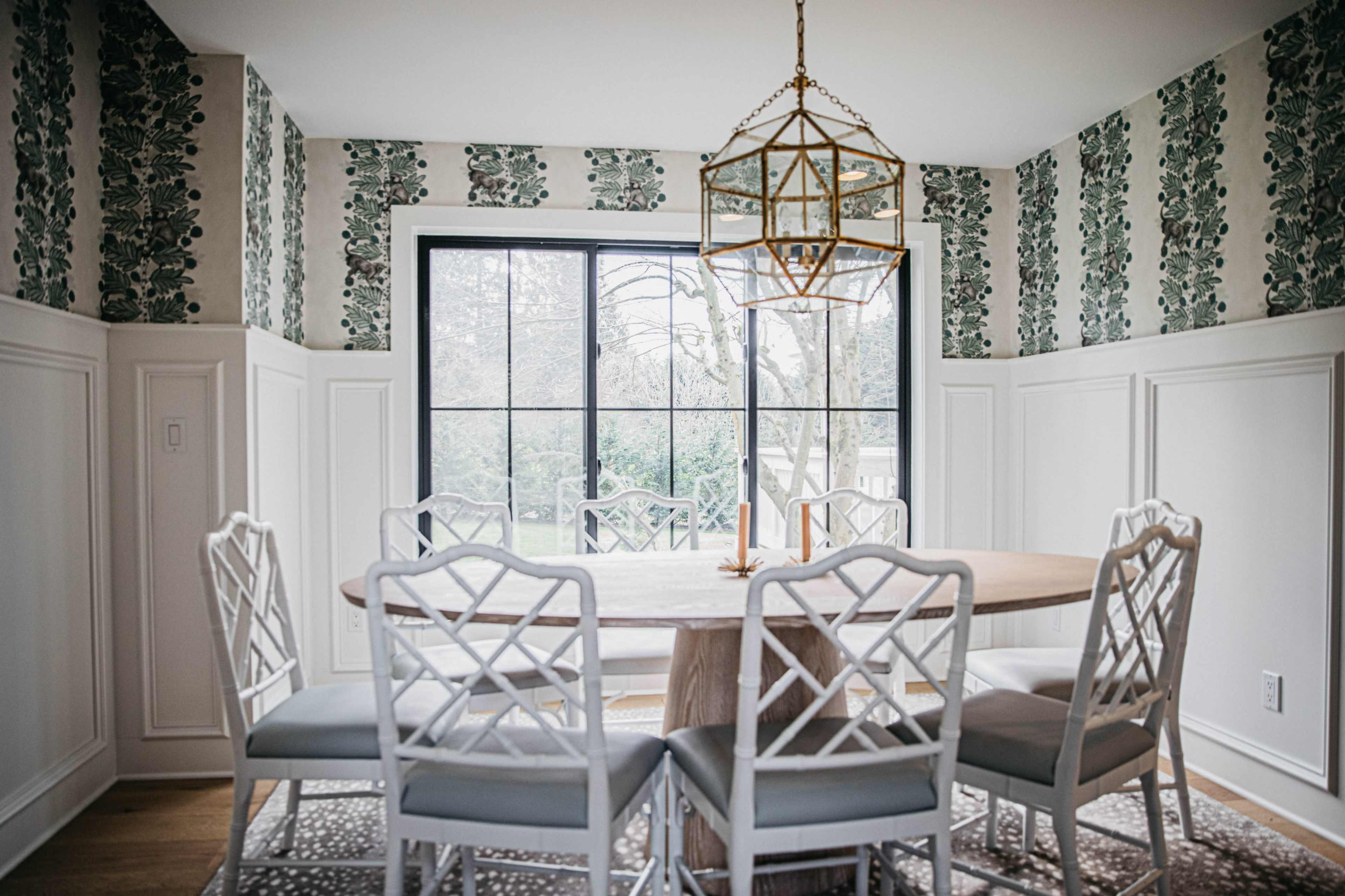 A bright dining room features a round wooden table surrounded by six white chairs, with patterned wallpaper and a large window overlooking a garden.