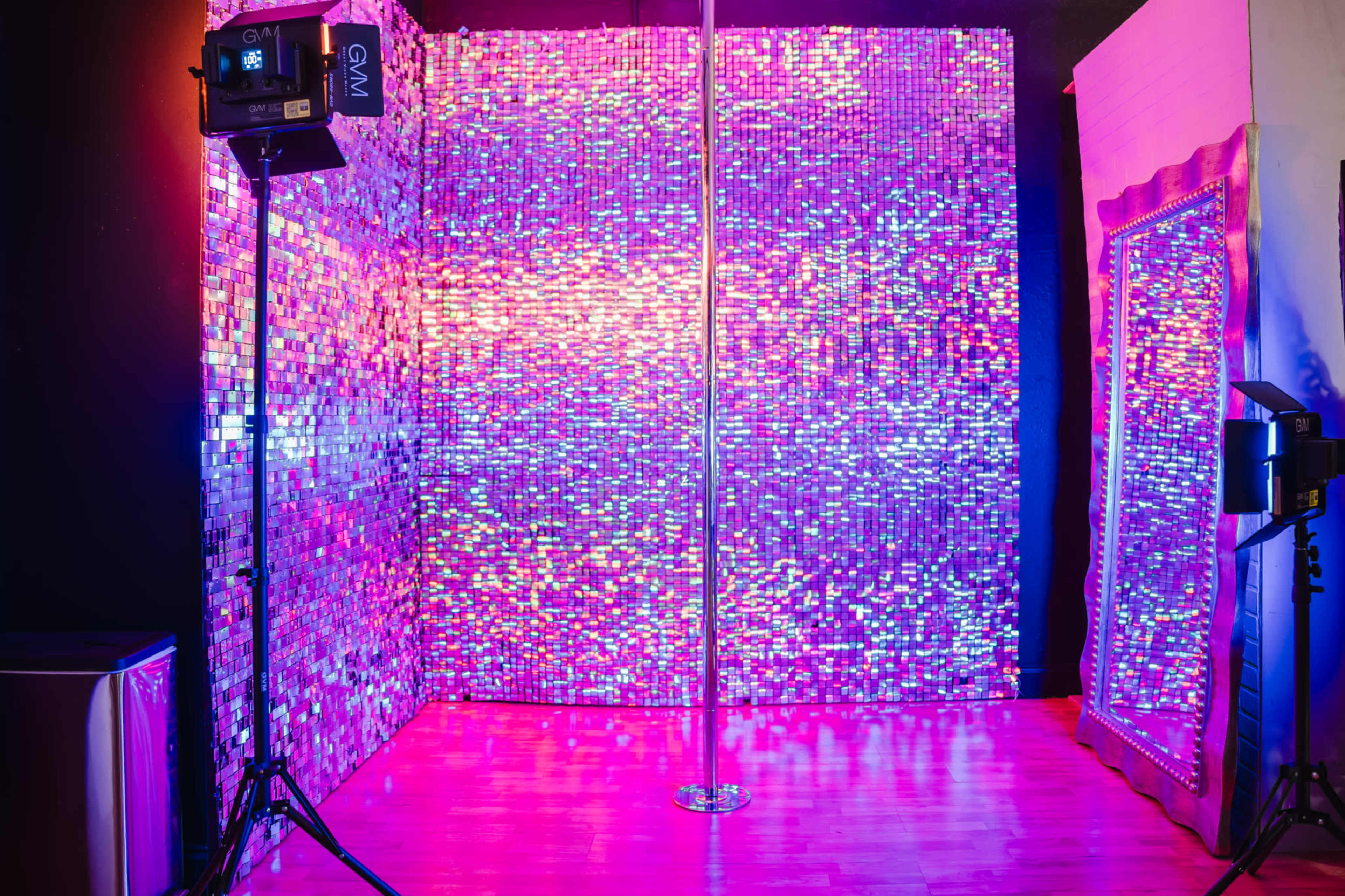 The image shows a brightly lit indoor space with a mirrored wall, a dance pole in the center, and colorful lighting effects throughout.