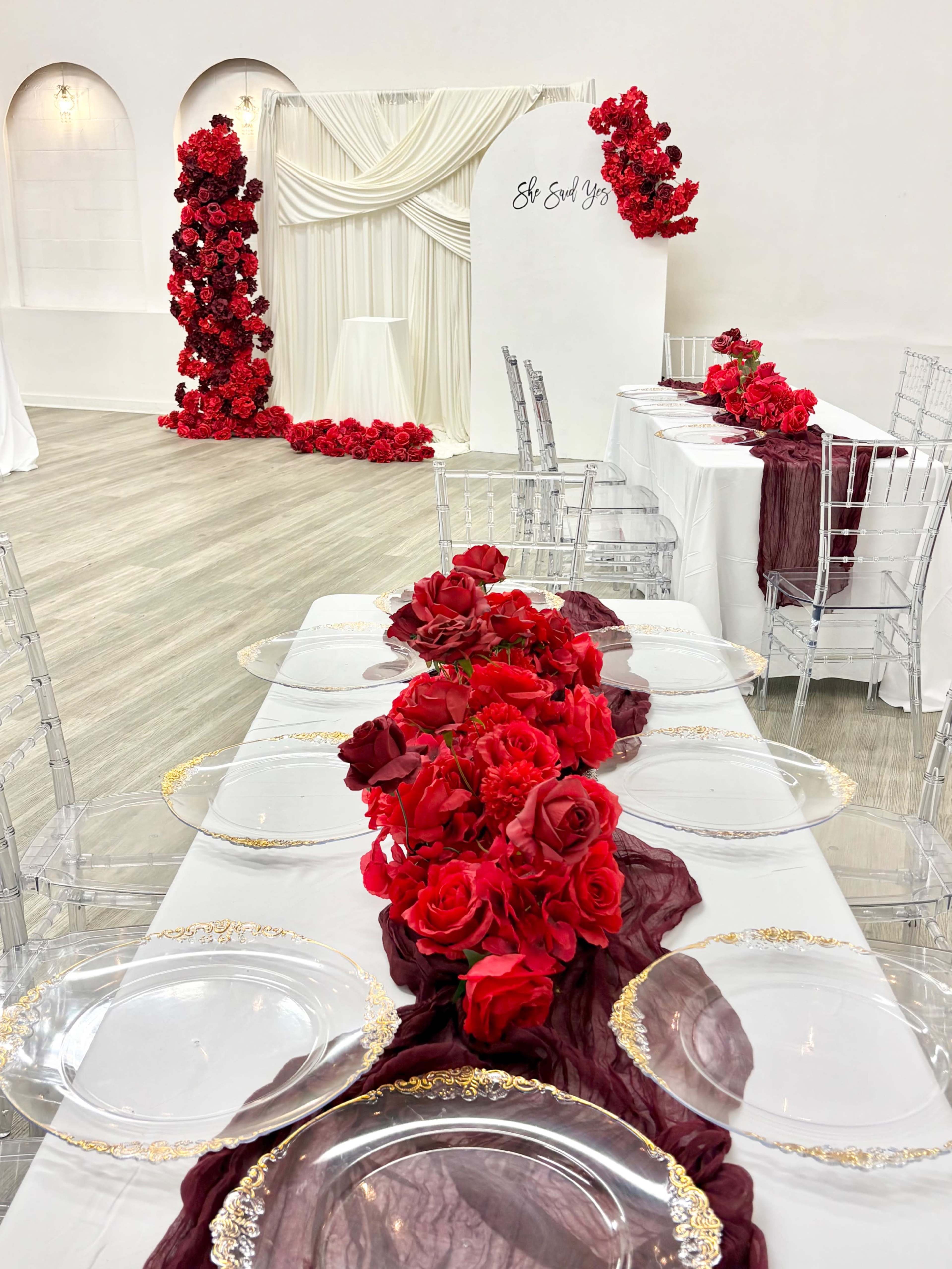 The image shows a decorated event space with white tables set with clear plates and red roses, accompanied by a backdrop of draped fabric and a floral arrangement.