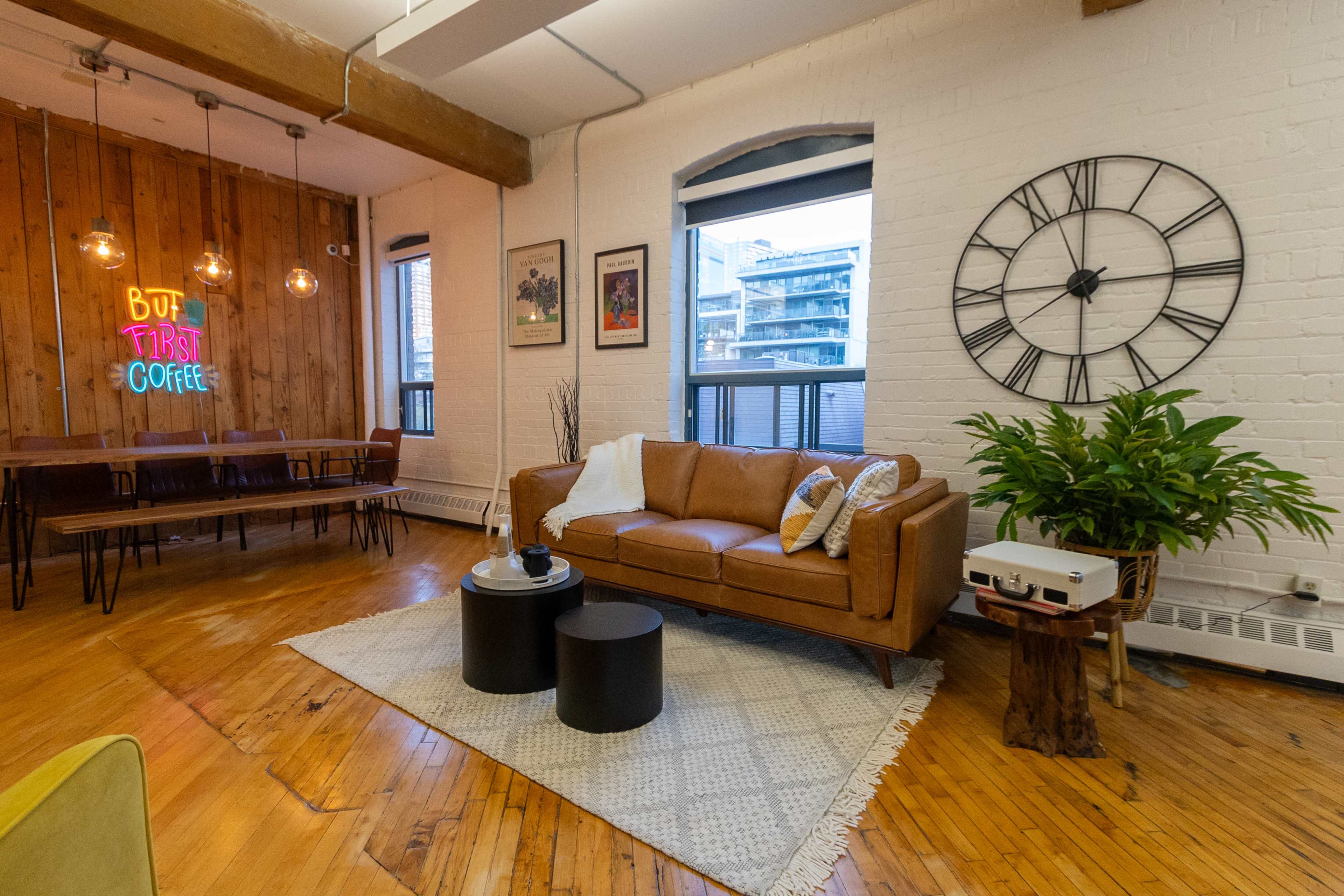 The image shows a cozy living area featuring a brown leather couch, a round coffee table, a large clock on the wall, and a neon sign that says "But First Coffee."