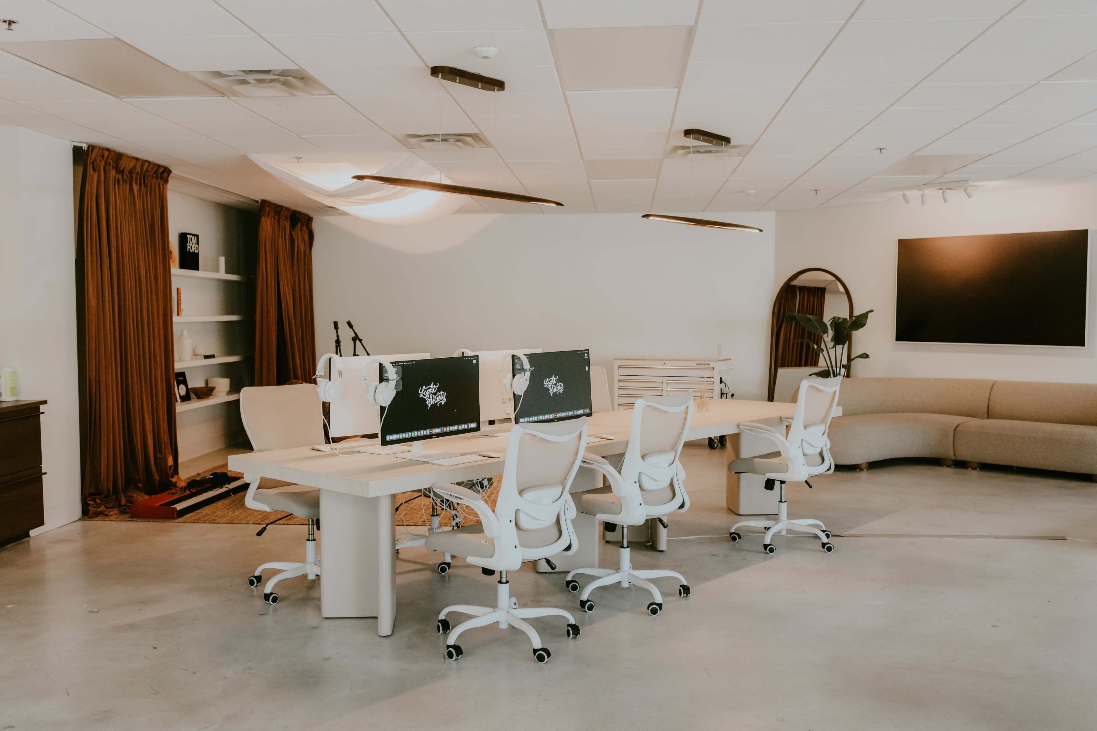 The image shows a modern office space with a long table, several white swivel chairs, and two computer monitors on the table, surrounded by minimalistic decor and a large sofa in the background.