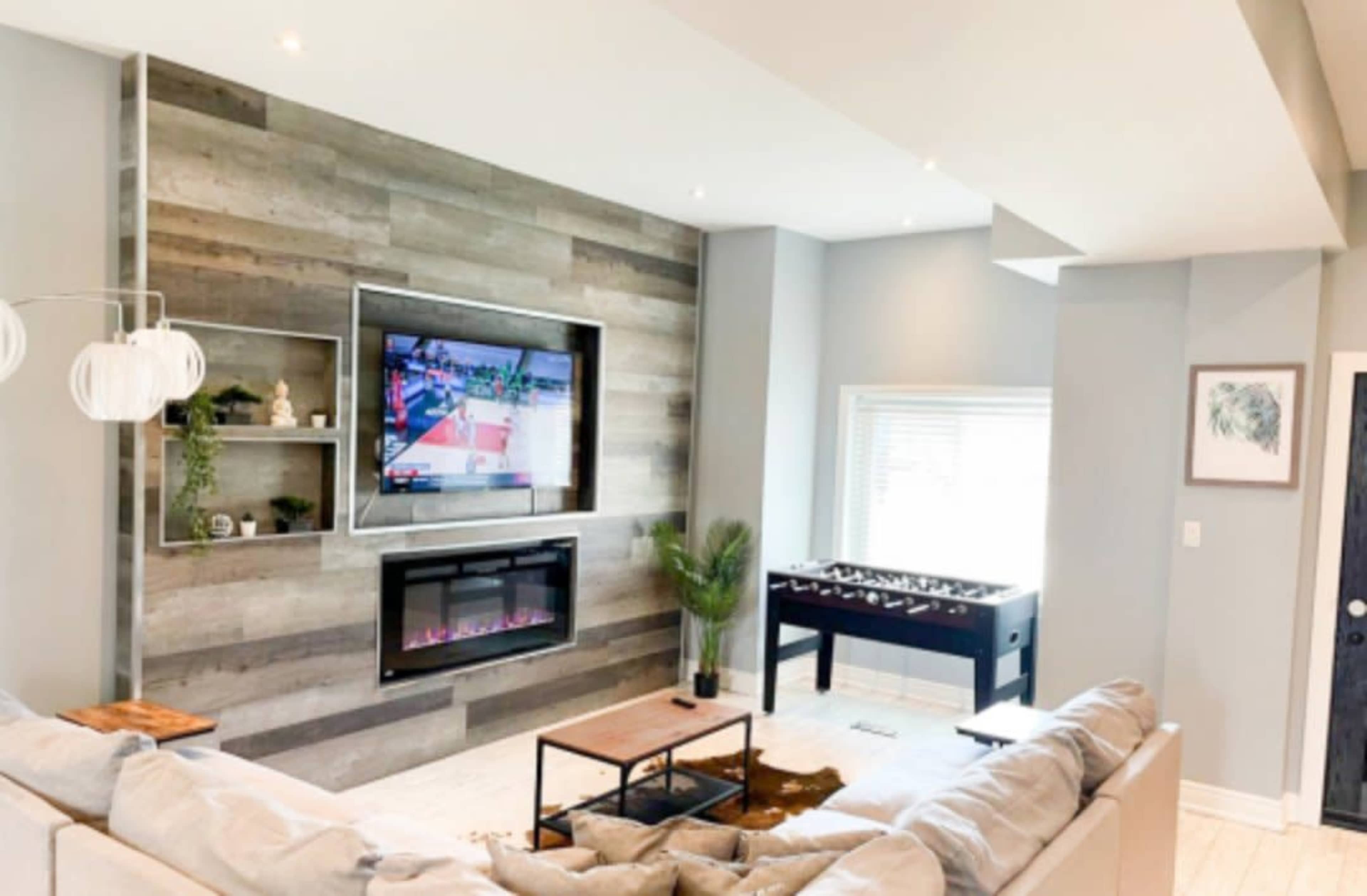 A modern living room features a wall-mounted TV above a sleek electric fireplace, flanked by decorative shelves, with a foosball table in the corner.