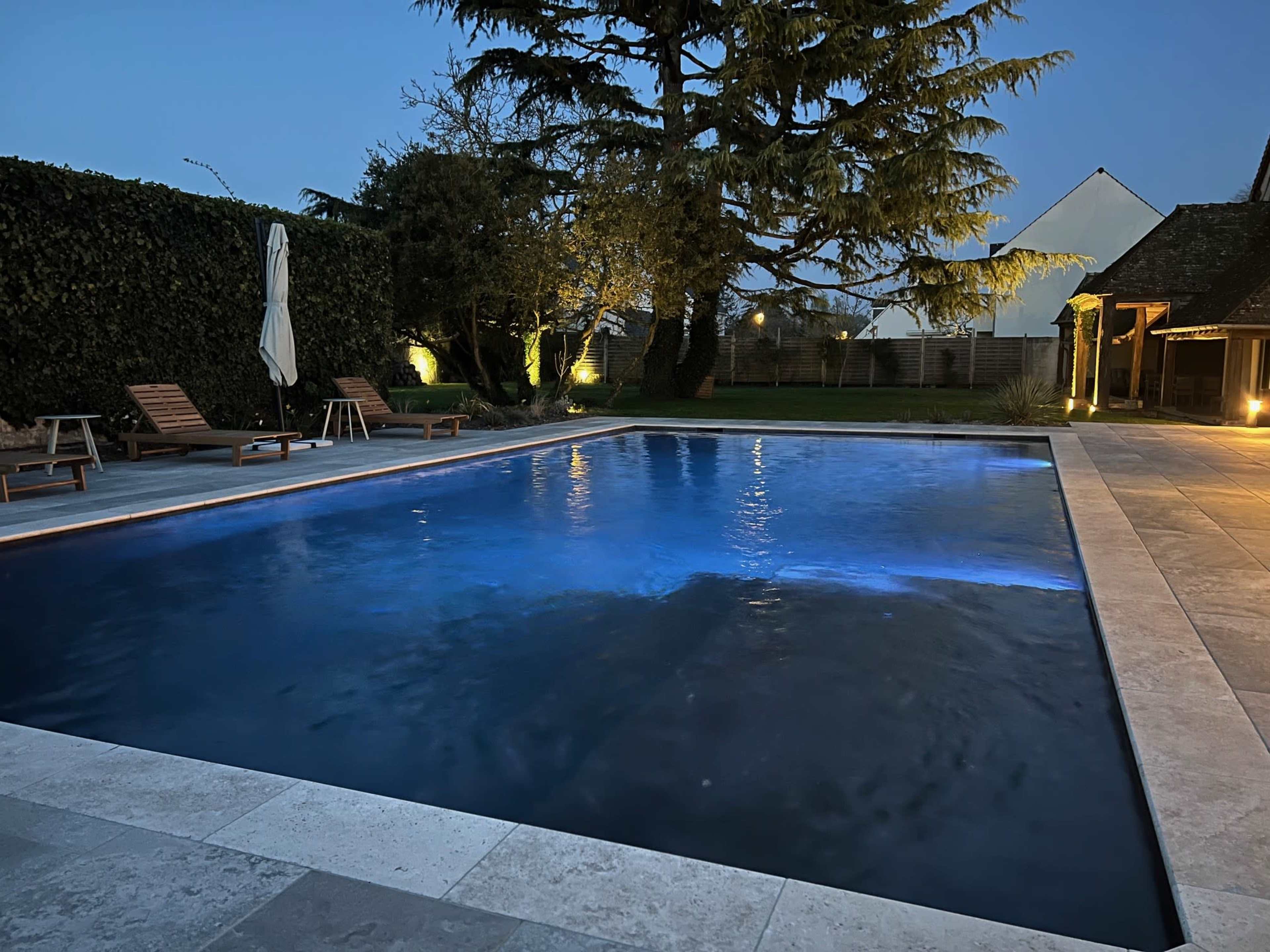The image shows a lit swimming pool surrounded by lounge chairs and a well-kept garden, with a house visible in the background.