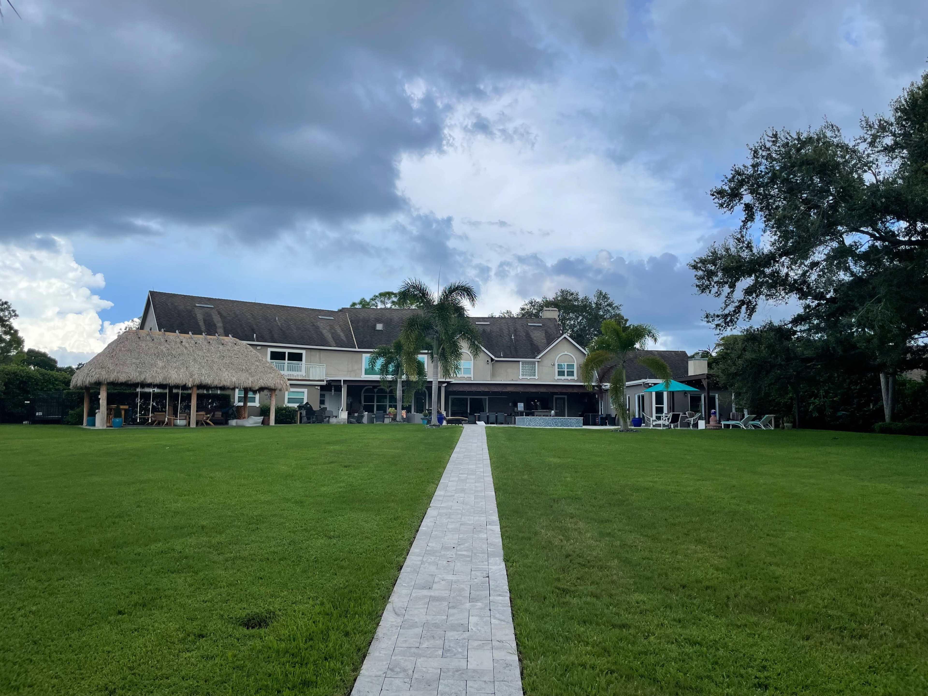 A large house with a thatched roof and a patio is set against a cloudy sky, facing a well-maintained lawn and a stone pathway.
