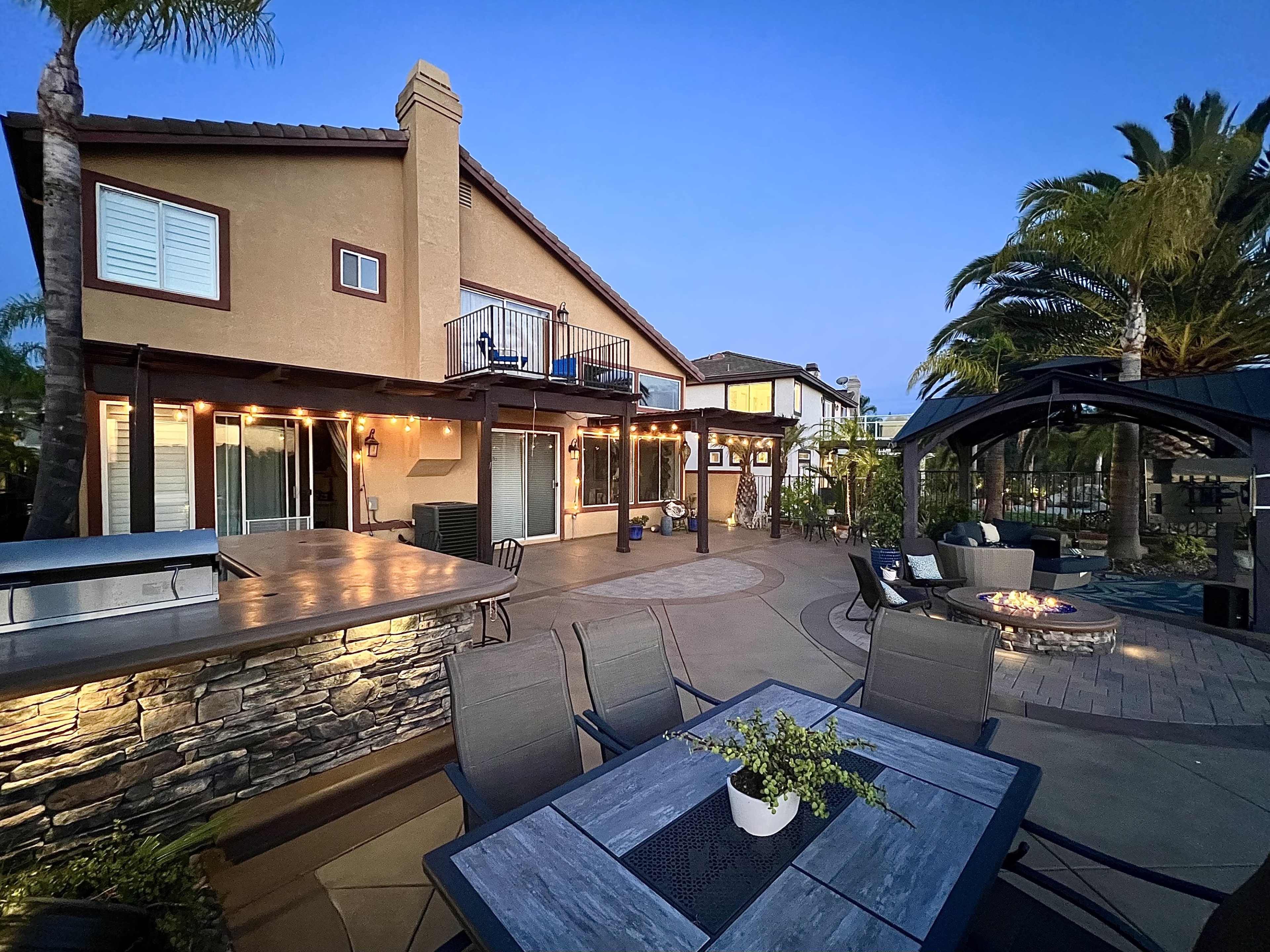 The image shows a spacious residential backyard with a patio area featuring a dining table, outdoor fire pit, and a house equipped with a balcony and palm trees in the background.