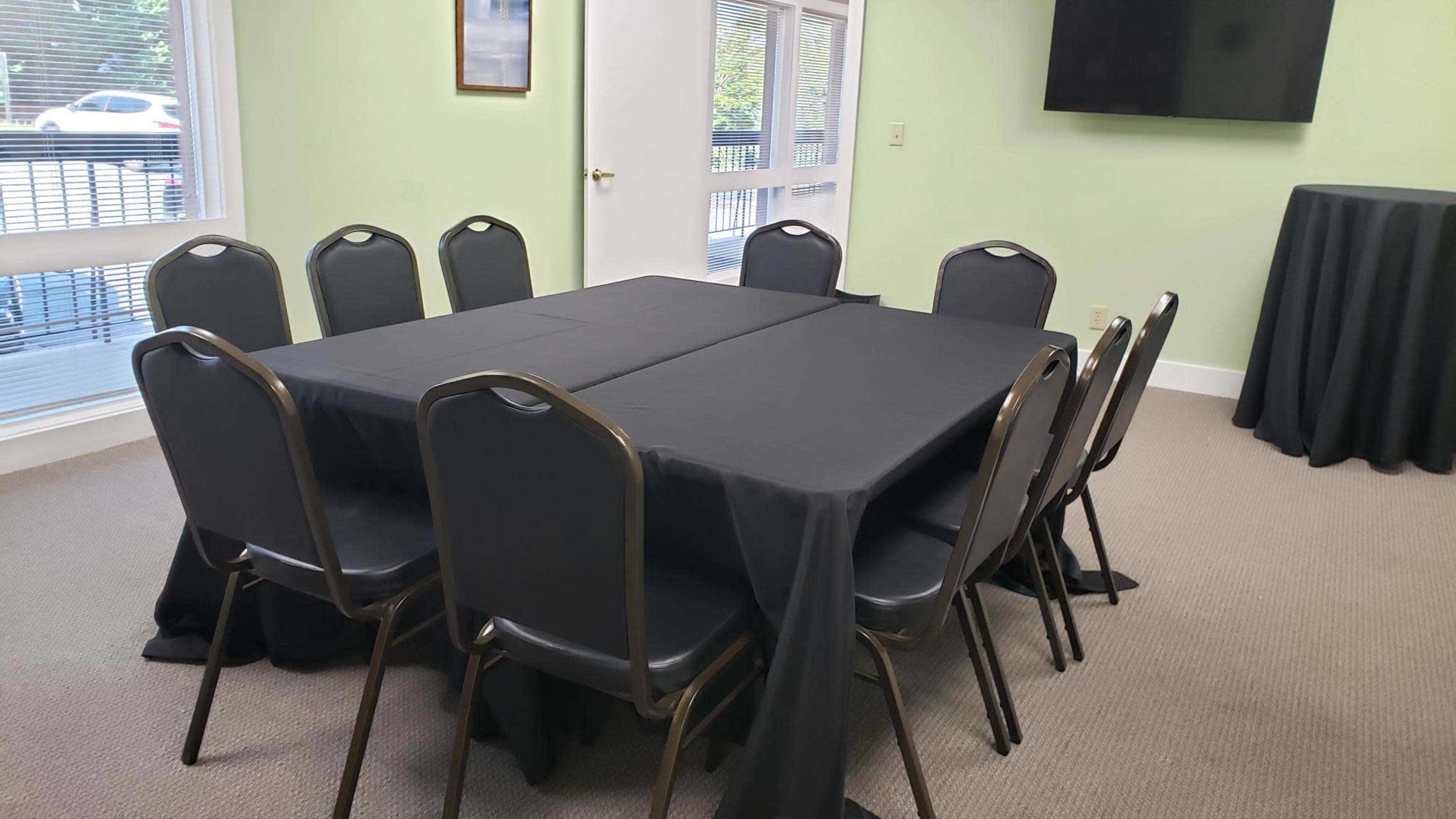 A rectangular table covered with a black tablecloth is surrounded by eight chairs in a small, light-colored meeting room.