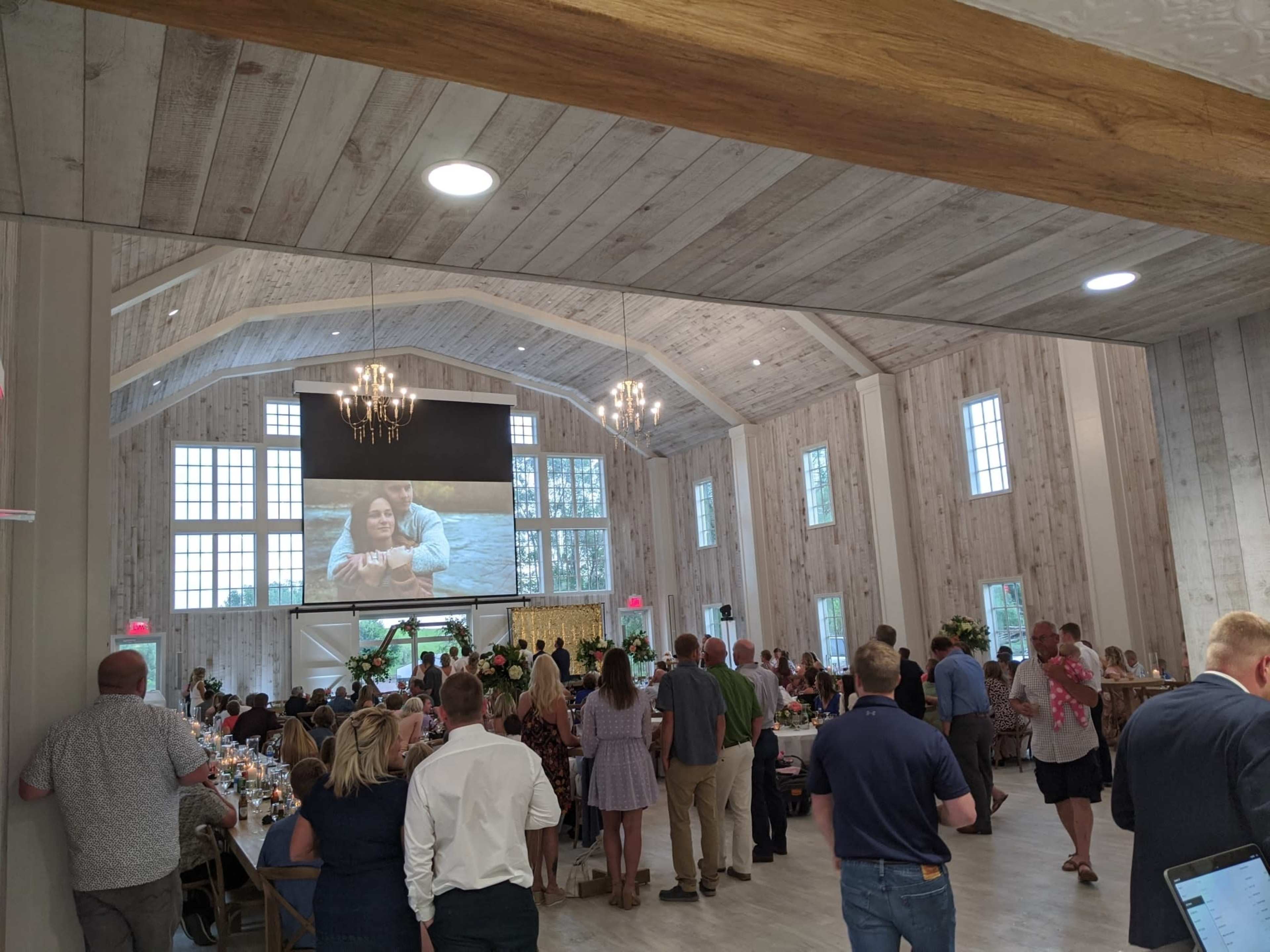 A large gathering is taking place in a spacious event hall, featuring long tables set for dinner and a large screen showing a couple, with guests mingling and seated around the room.