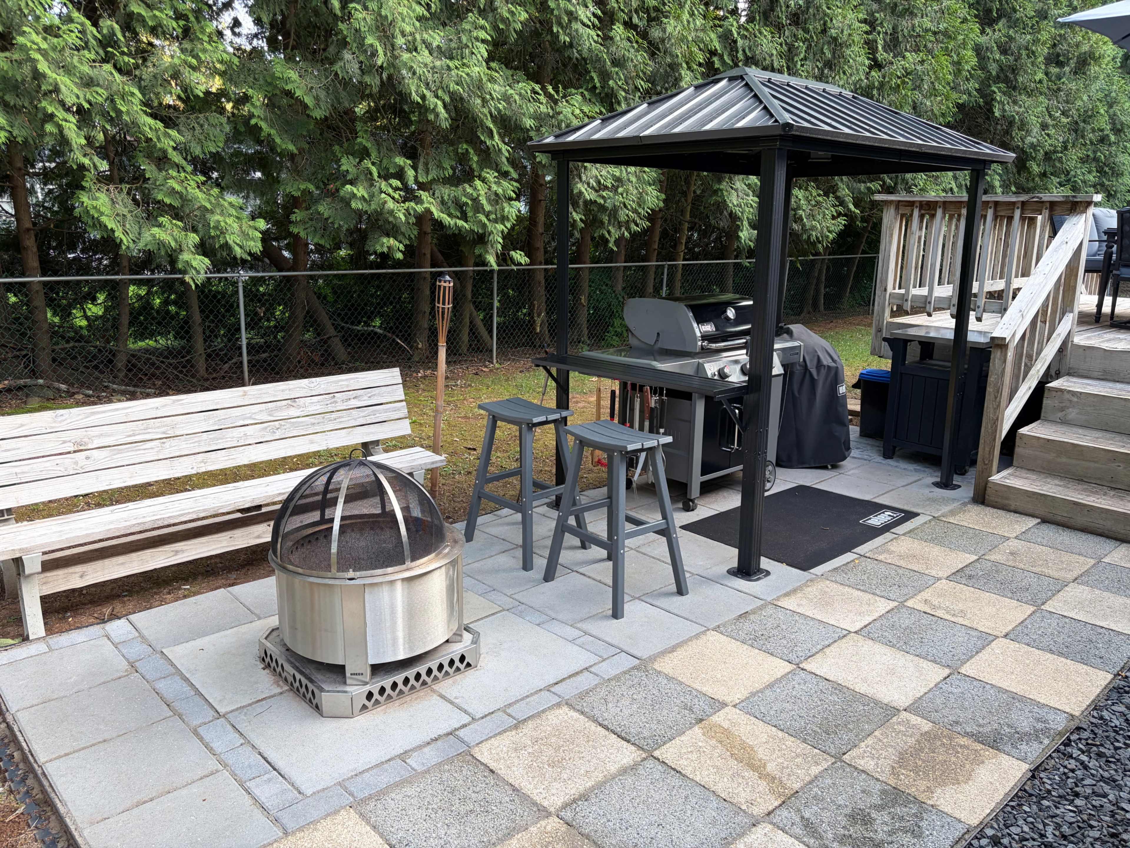The image shows an outdoor grilling area with a gazebo, a grill, a fire pit, a wooden bench, and stone paving.