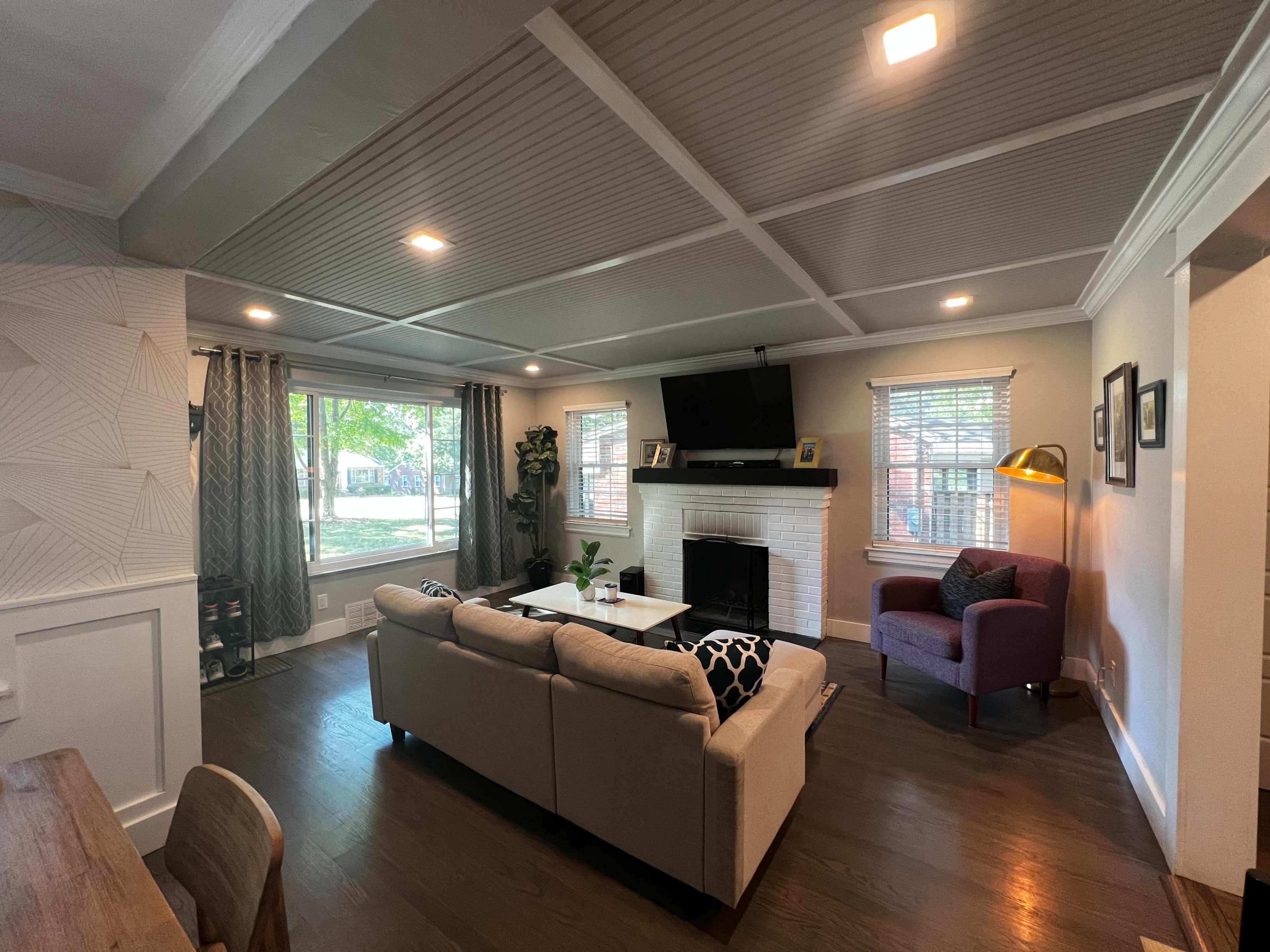 A living room with a beige sectional sofa, a purple armchair, a coffee table, and a television mounted above a white fireplace, accented by large windows letting in natural light.