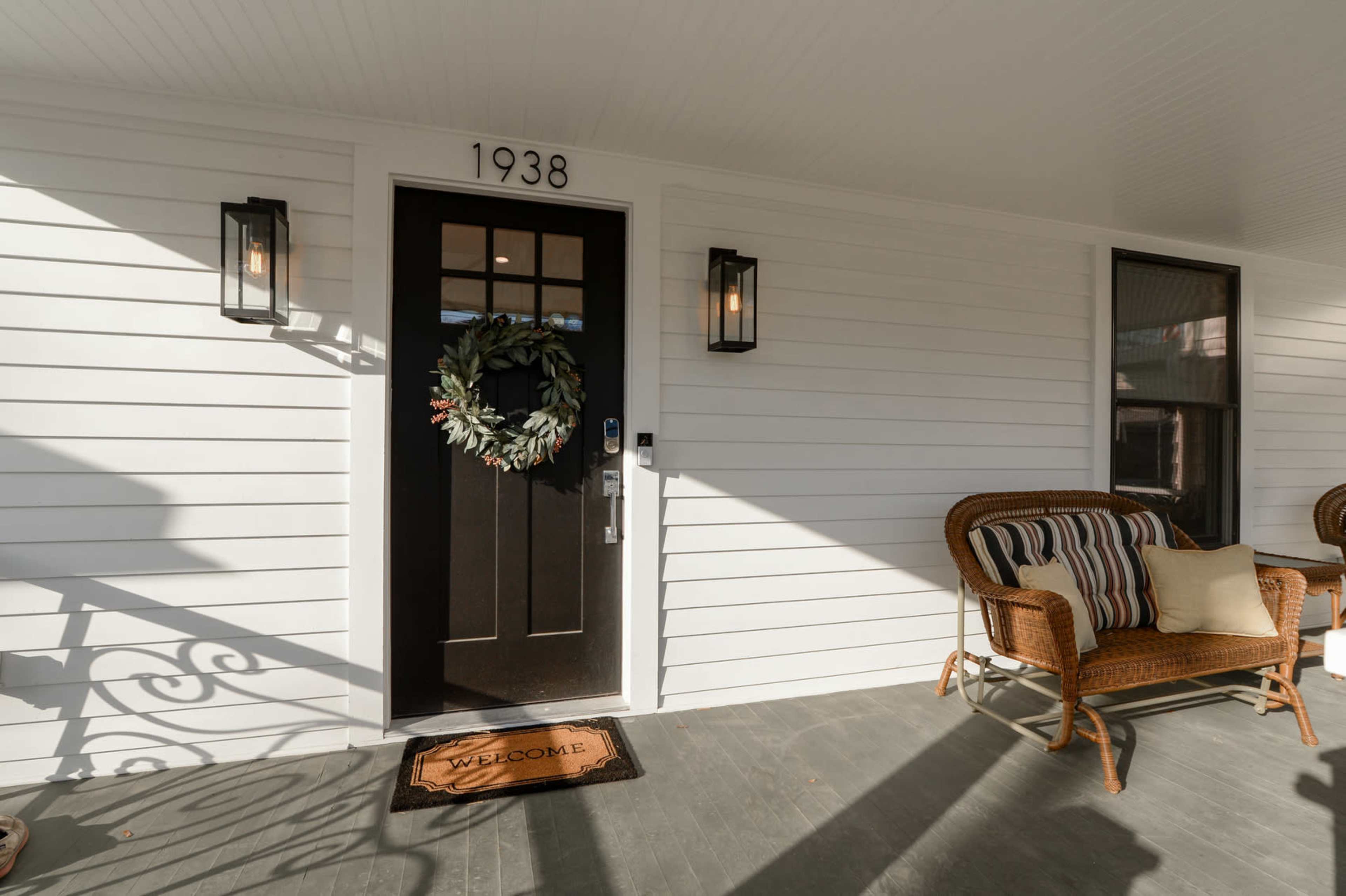 A front porch featuring a dark door with a wreath, two wall sconces, and a welcome mat in front of a wicker chair set against a white-paneled wall.