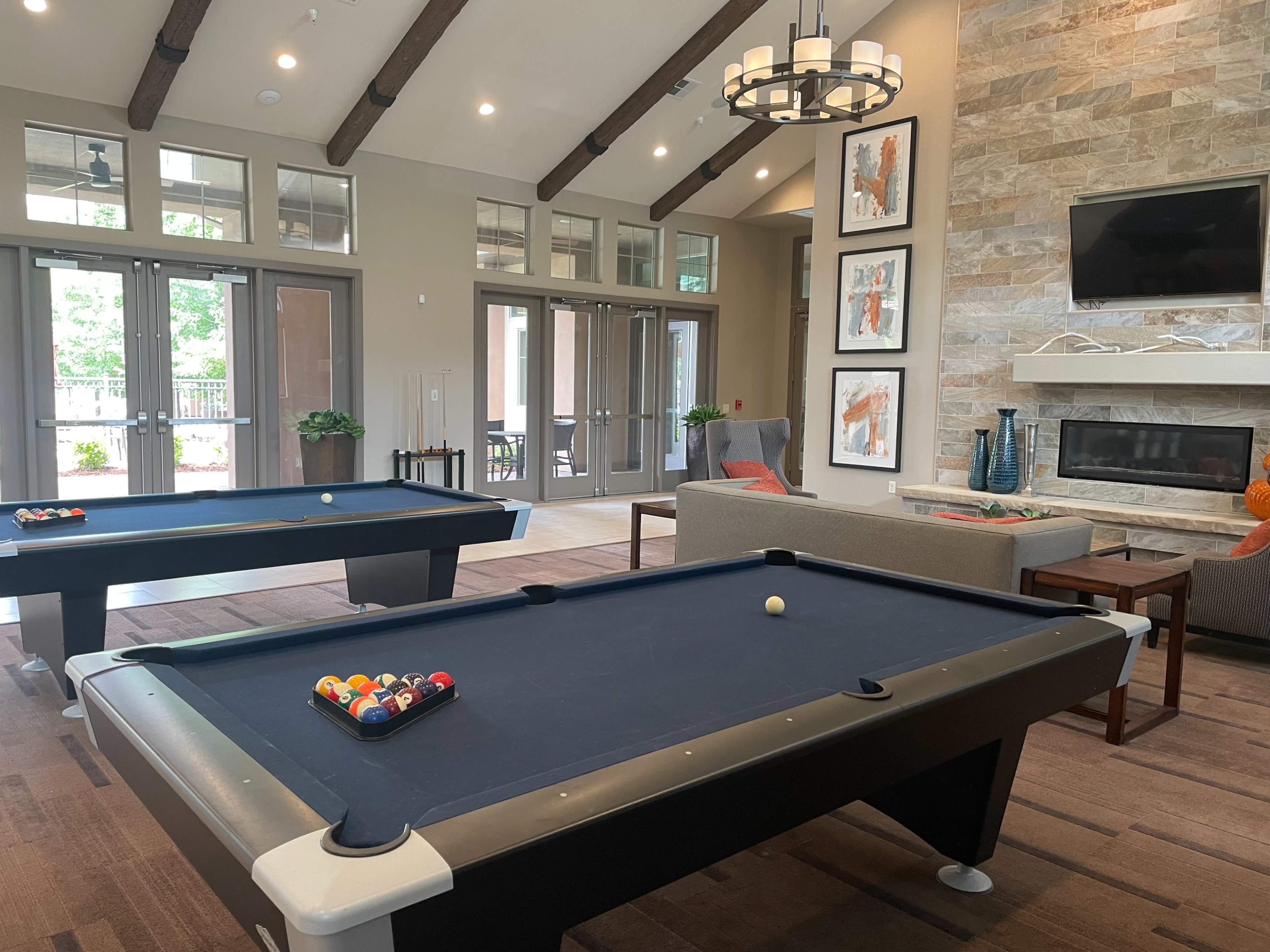 The image shows a spacious common area featuring two pool tables, a seating arrangement, and a stone fireplace with large windows allowing natural light.
