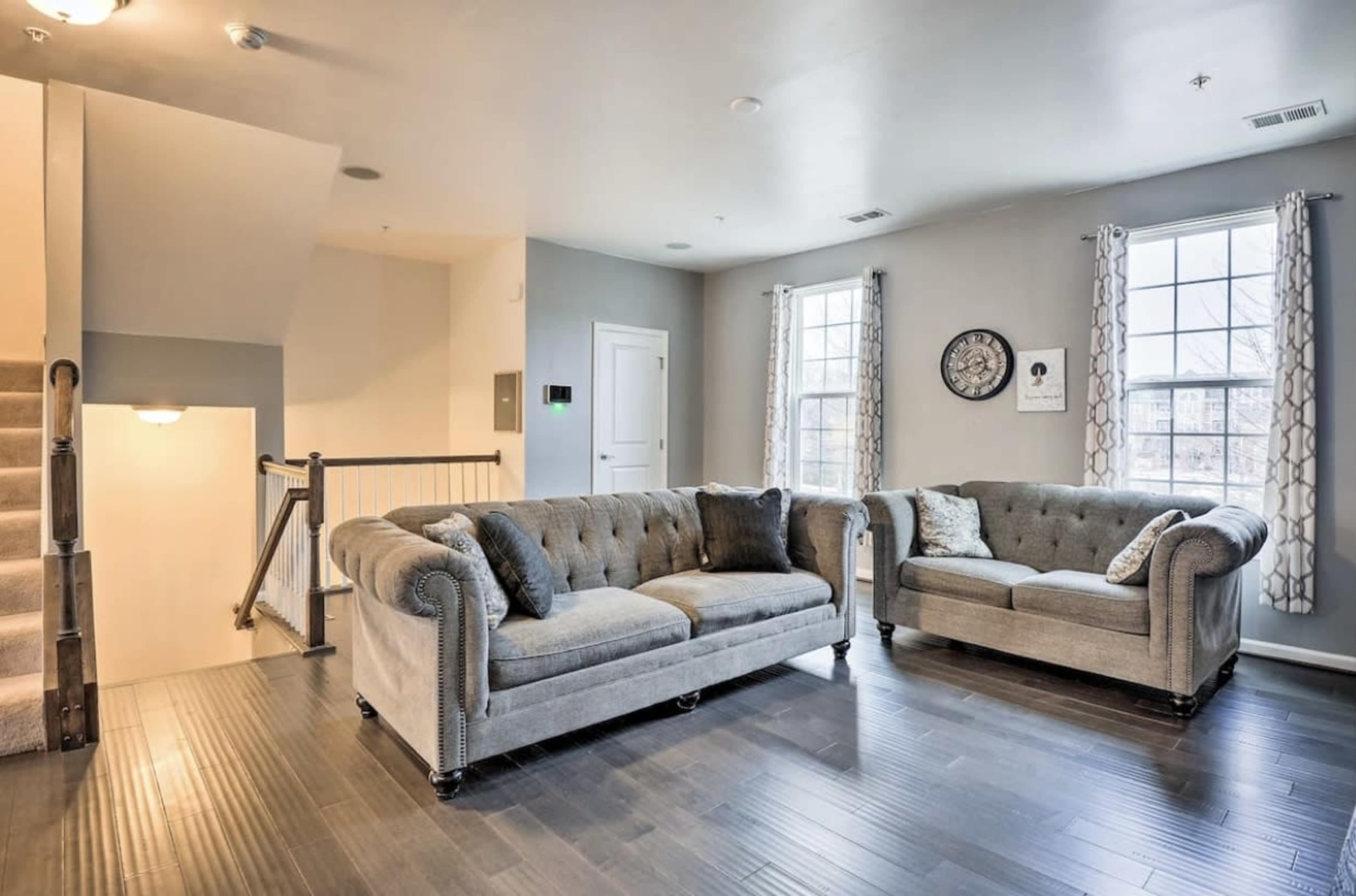 A living room with two grey tufted sofas, a staircase in the background, and large windows with decorative curtains.