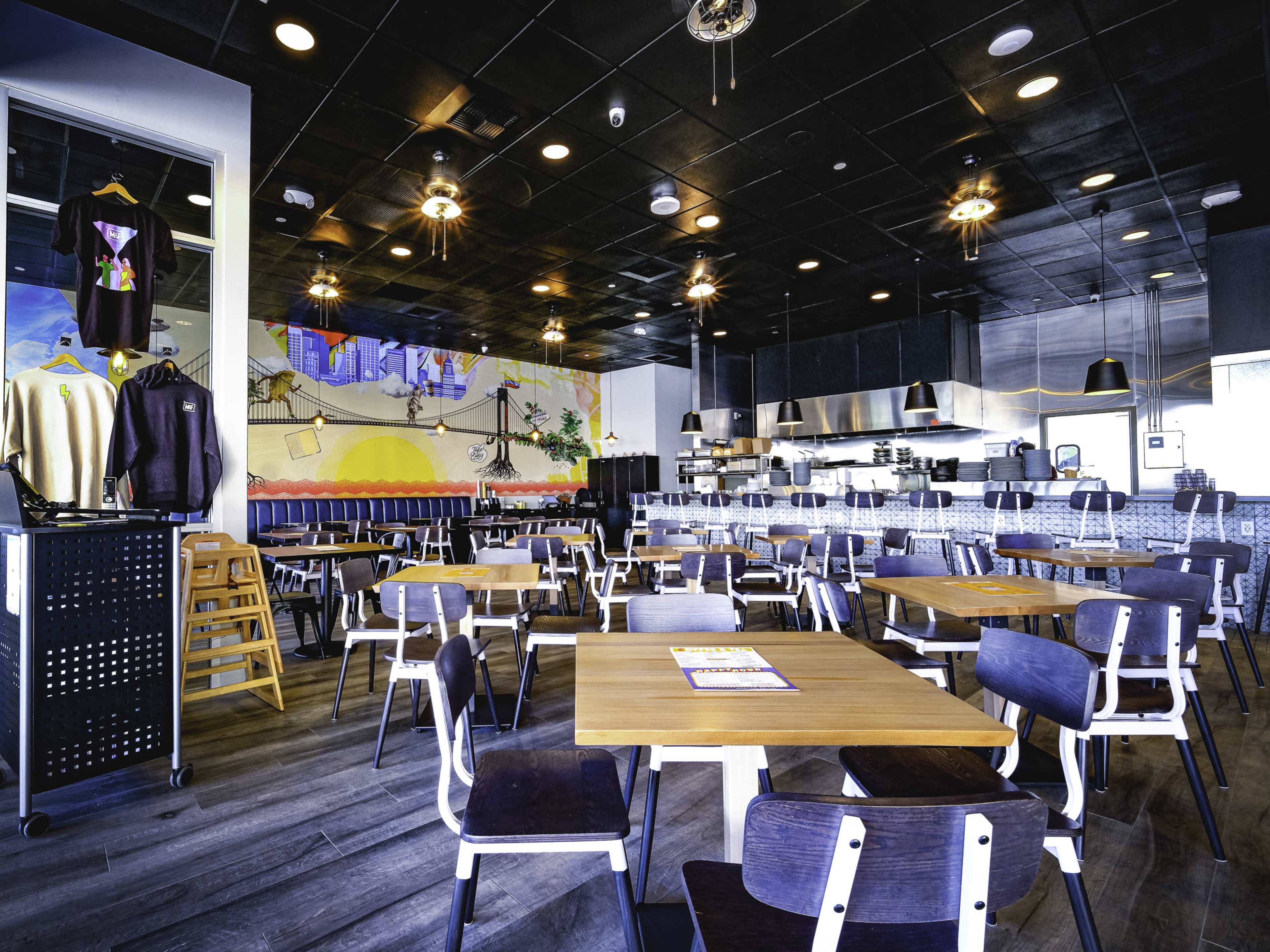 The image shows a modern restaurant interior with several wooden tables and chairs, a large mural on one wall, and kitchen equipment visible in the background.