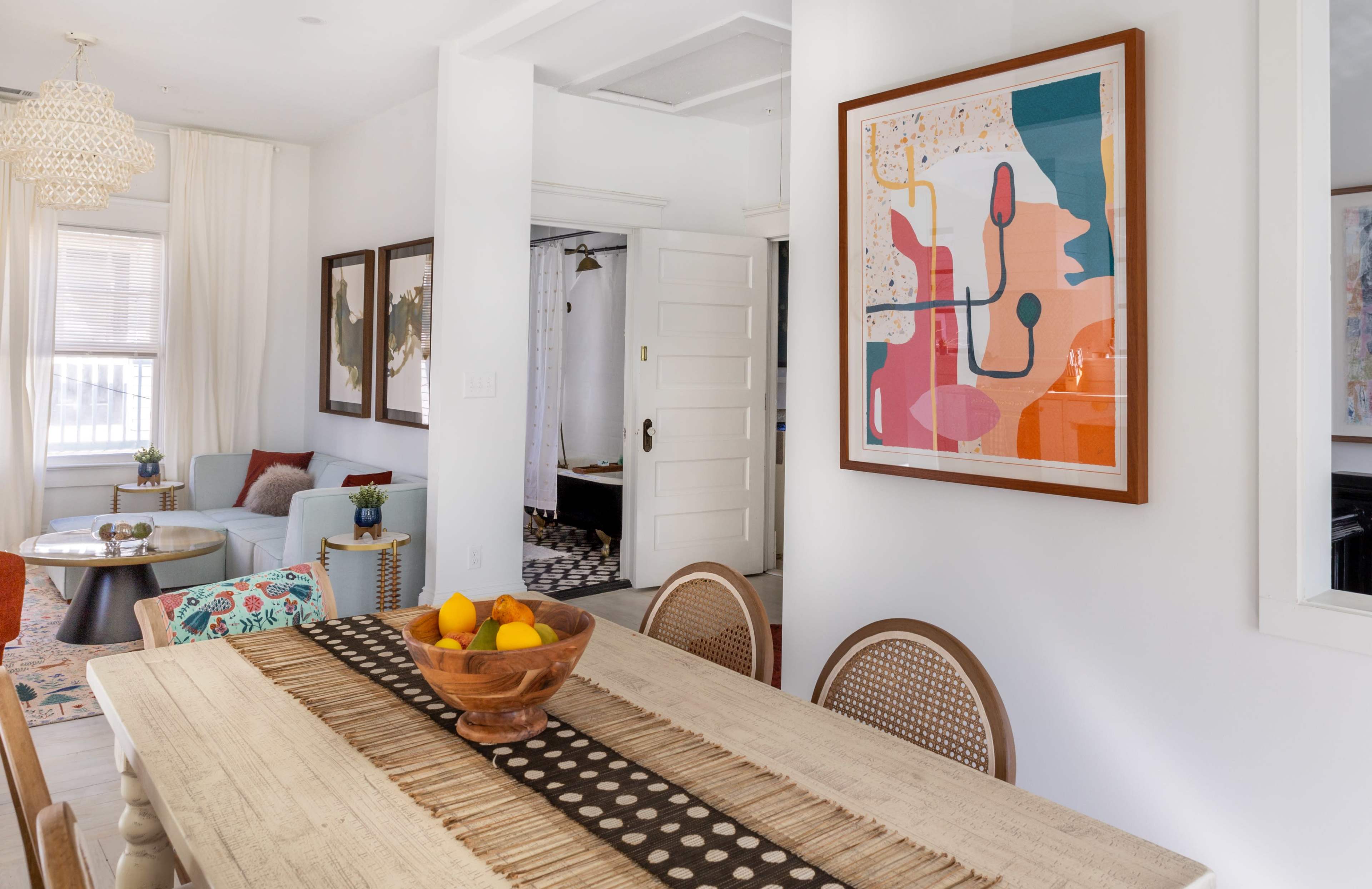 A dining table with a bowl of fruit is set in a bright, modern dining area adorned with framed artwork and a cozy seating arrangement.