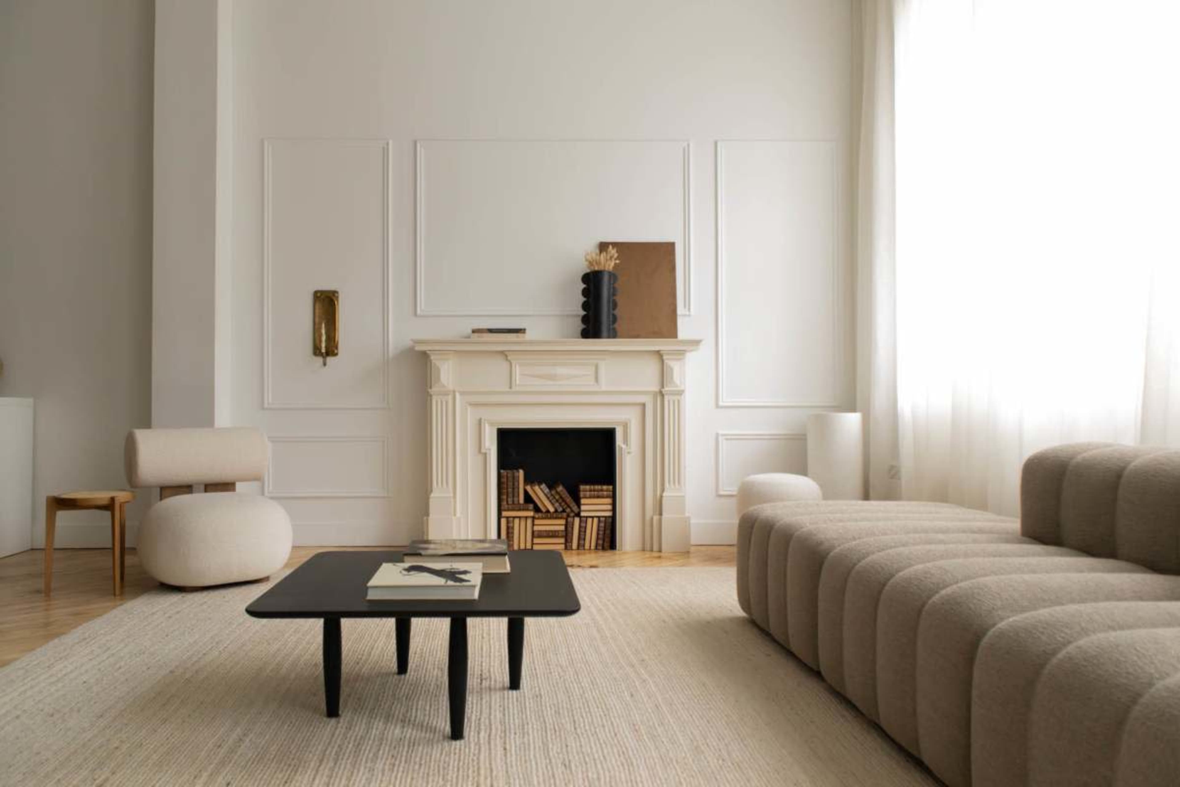 The image shows a modern living room featuring a beige couch, a black coffee table, a fireplace with books stacked inside, and soft natural light filtering through sheer curtains.