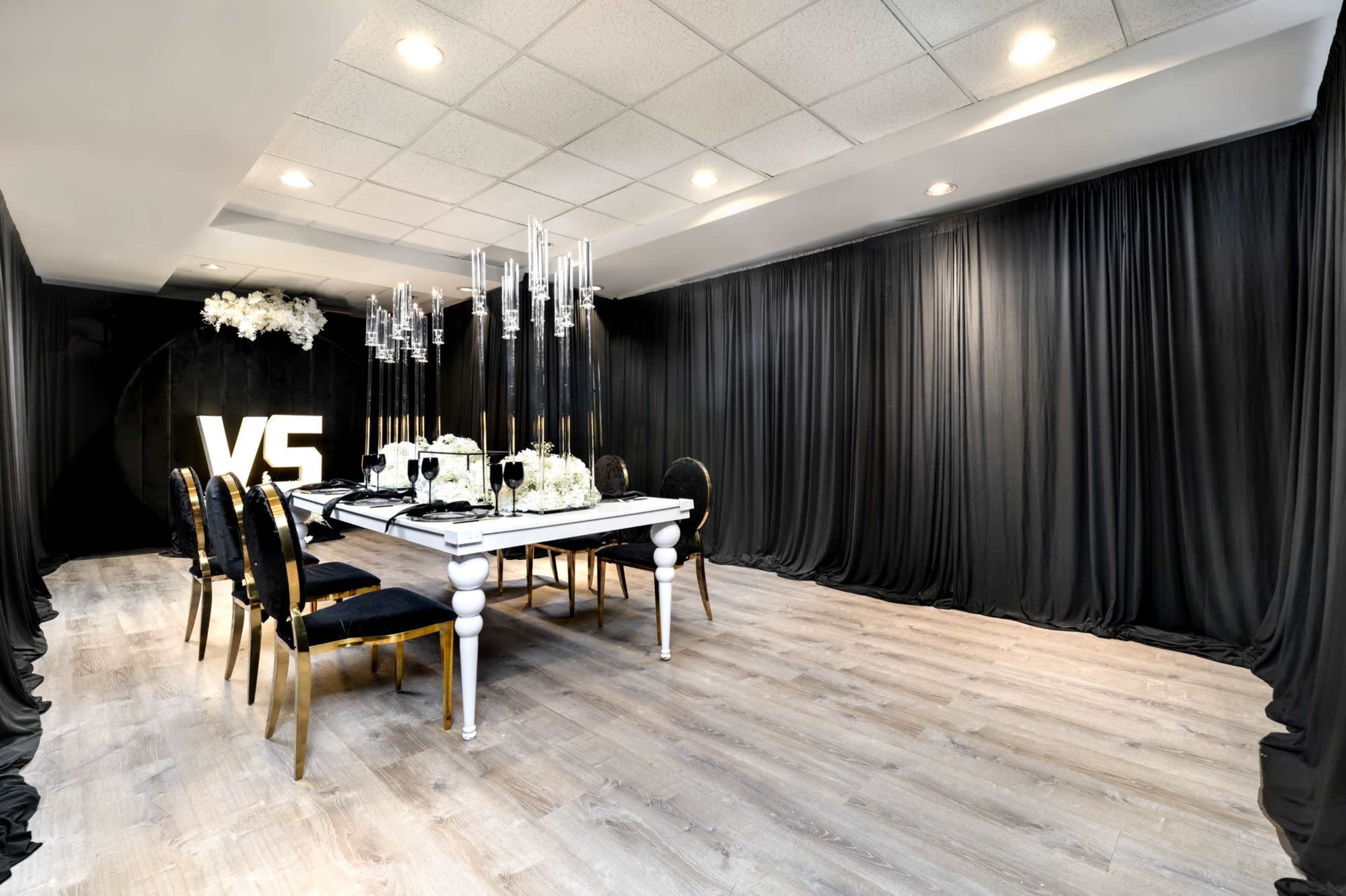 A dining room with a long white table set for a meal, surrounded by black upholstered chairs, with black drapery and large light fixtures overhead.