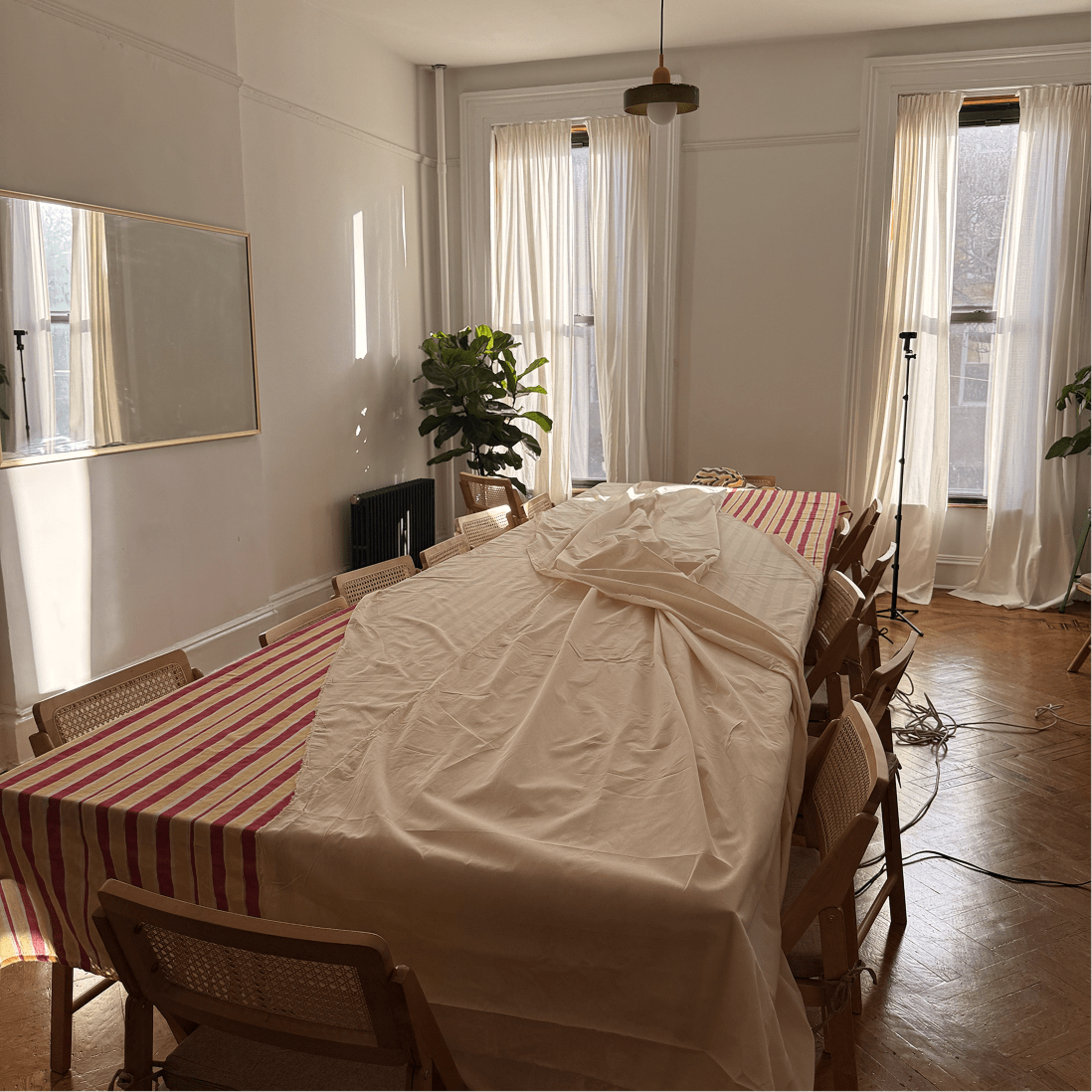 A long table covered with a white cloth is set up in a bright room with large windows, surrounded by wooden chairs and greenery.
