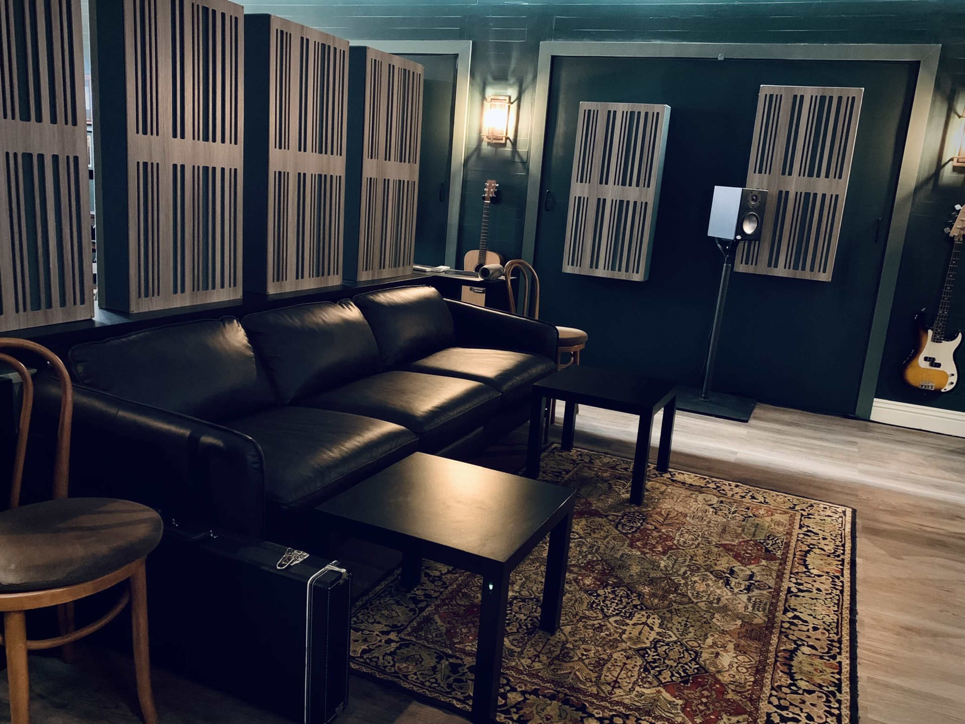 A music studio interior featuring a black leather sofa, two small tables, soundproofing panels on the walls, and musical instruments displayed nearby.